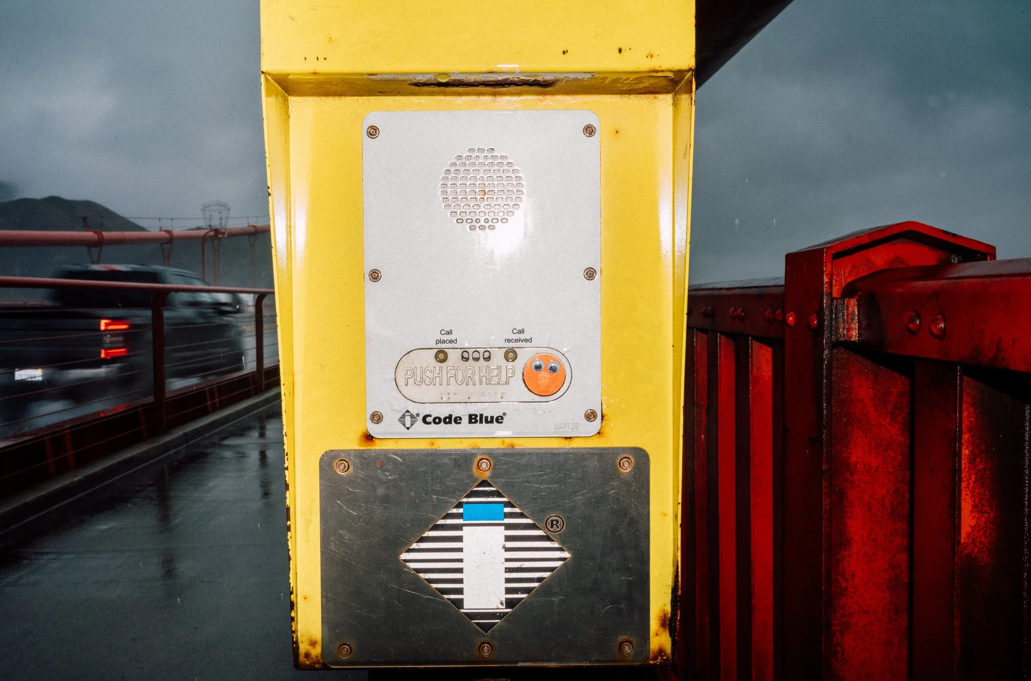 A yellow toll booth with a white speaker and a call for help button, located on a wet bridge with moving cars in the background and a cloudy sky.