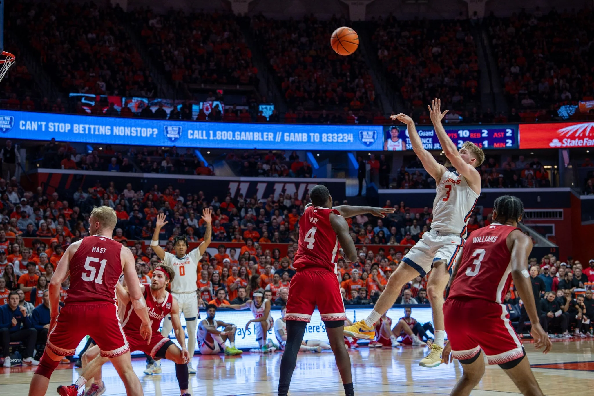 A basketball game in progress at a stadium with players jumping and reaching for the ball, including players in white and red uniforms, with fans in the background and digital banners around the arena.