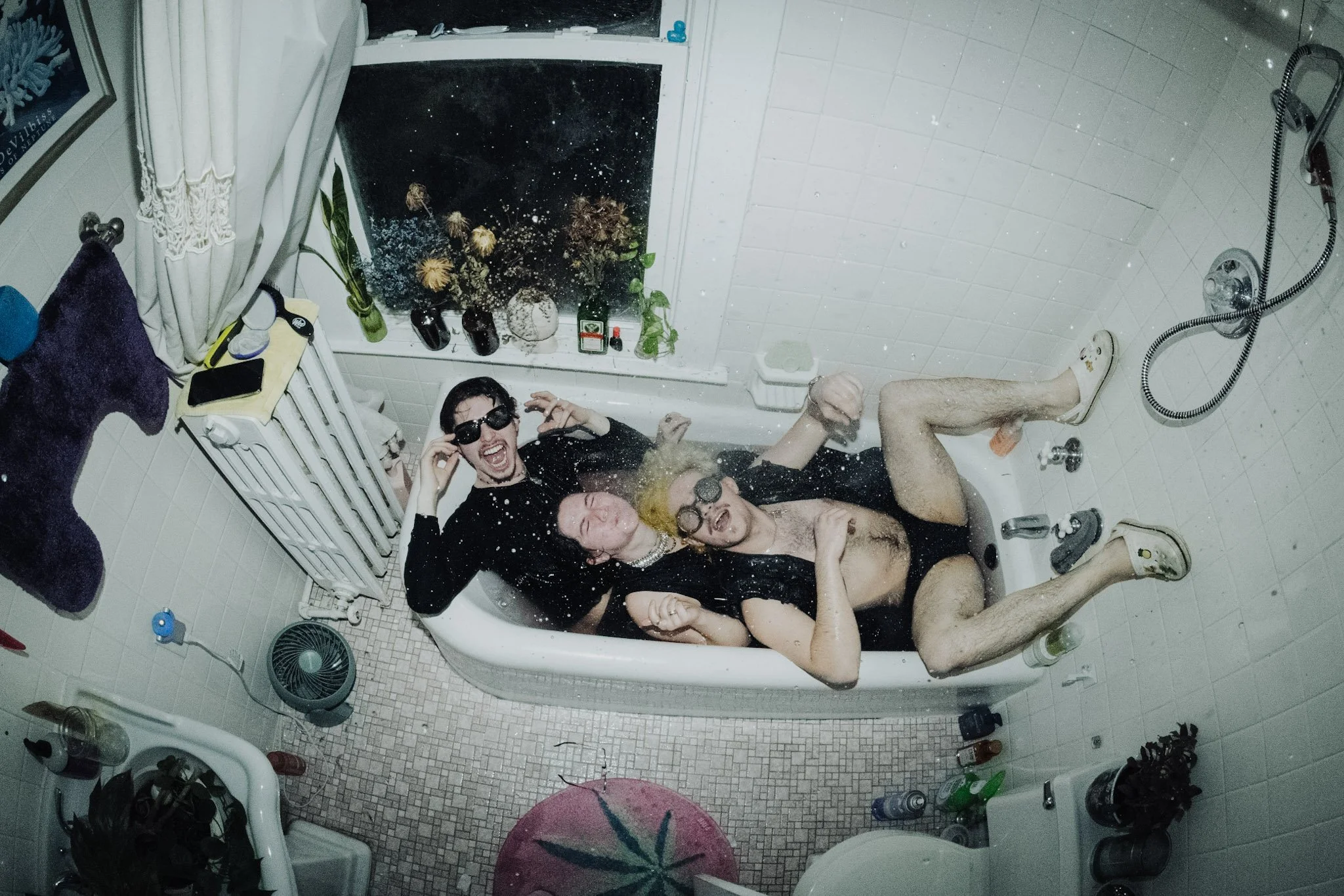 Three people in a bathtub taking a selfie, with water splashing around. They are wearing sunglasses and appear to be having fun.