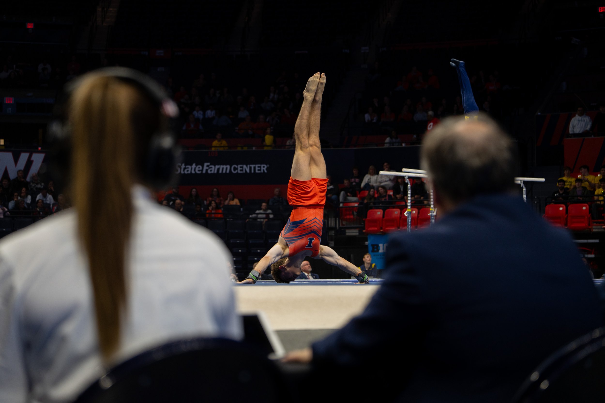 A male gymnast in a red and blue uniform performs a handstand on the balance beam at an indoor sports venue, watched by two judges seated in front of him.