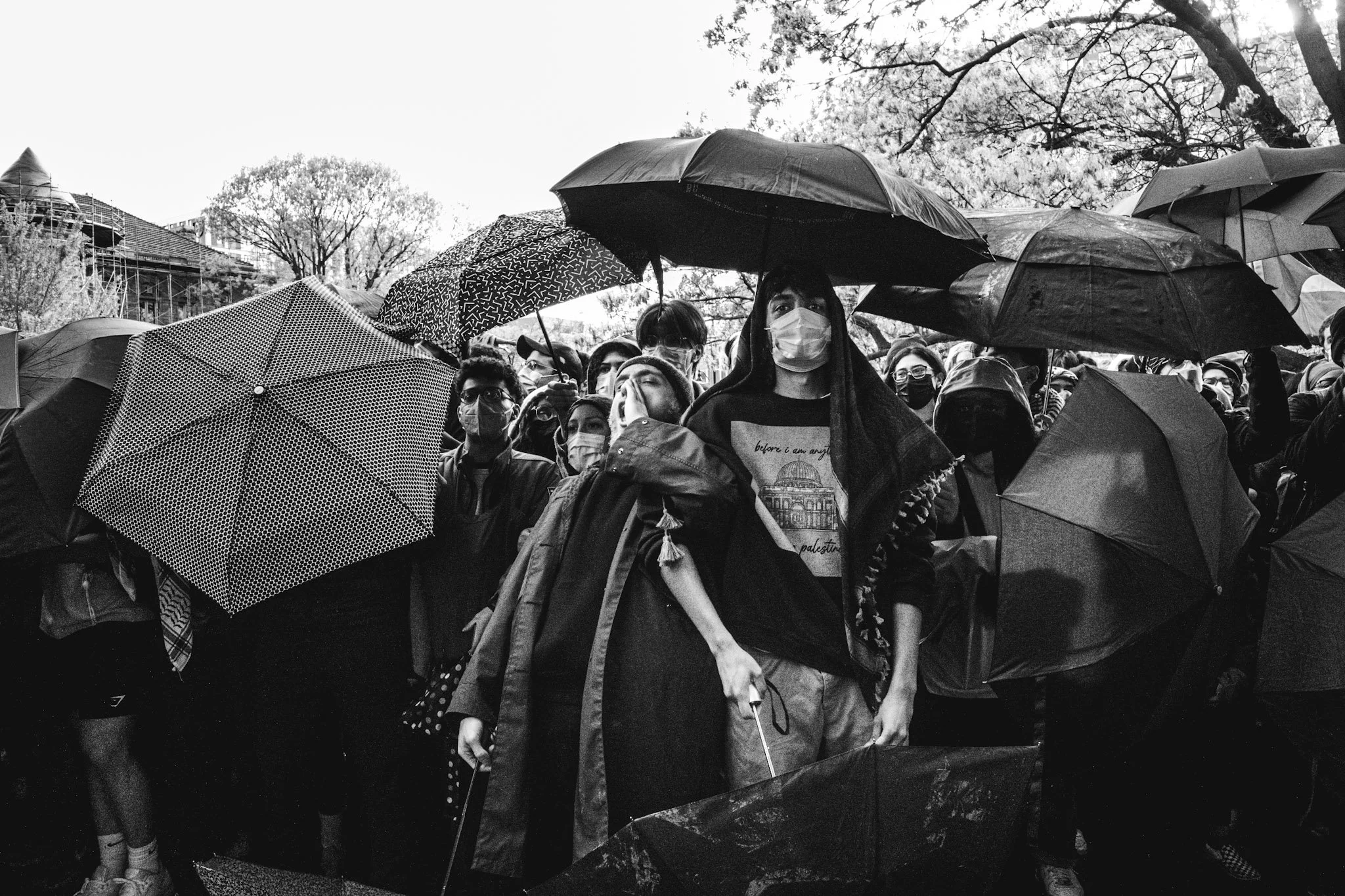 A crowd of people wearing face masks under umbrellas during rain, gathered outdoors likely for a protest or gathering.