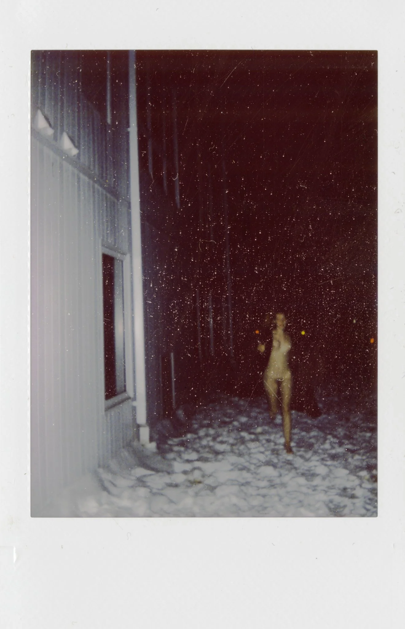 A Polaroid picture showing a nude mannequin walking outside in snowy weather at night, near a building with a window, with snow on the ground and visible snowflakes falling.