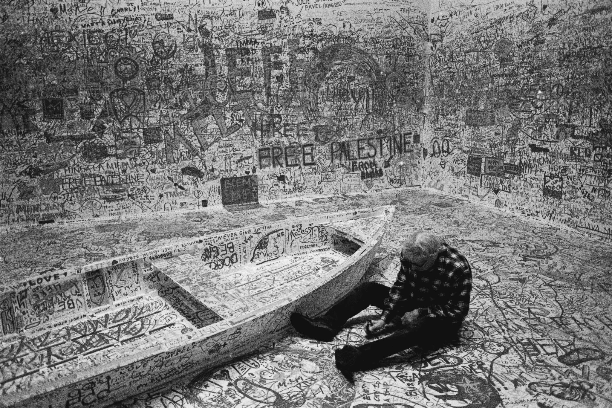 An elderly man sits on a floor covered in graffiti and sketches in a room with graffiti-covered walls and a boat-shaped structure in the middle, also covered in graffiti.