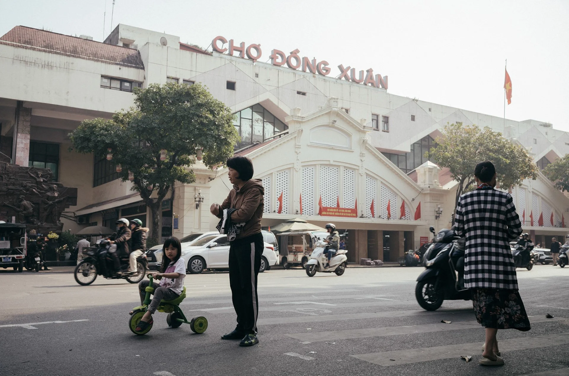 People on the street in front of a building with a sign that reads 'Chợ Đông Xuân'; a woman with a child on a tricycle, a woman walking, motorcycles passing by, and trees lining the street.