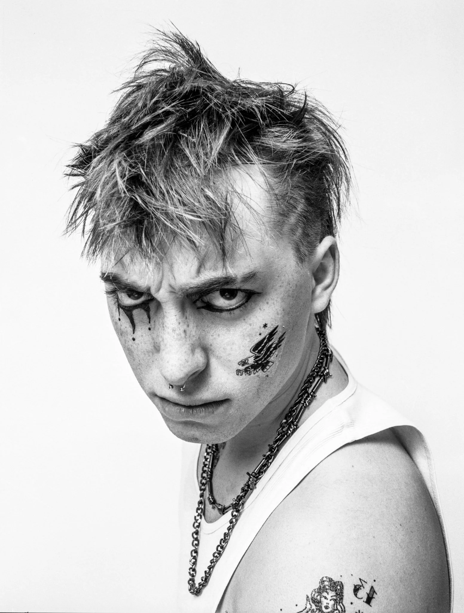 Black and white portrait of a person with spiky hair, tattoos on the face and arm, nose piercings, intense expression, wearing chains and a sleeveless shirt.