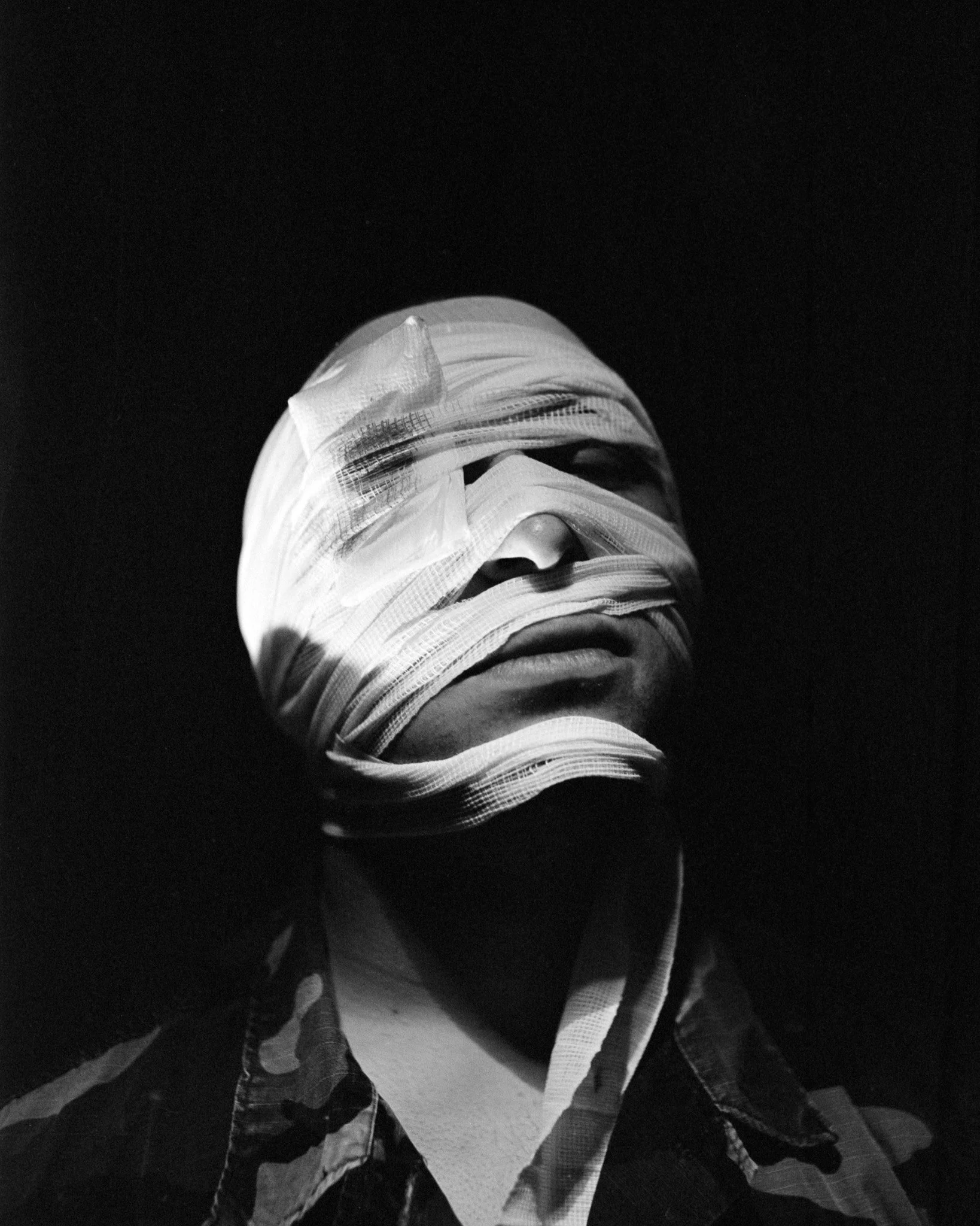 A person with their face wrapped in bandages, appearing in black and white with a dark background.