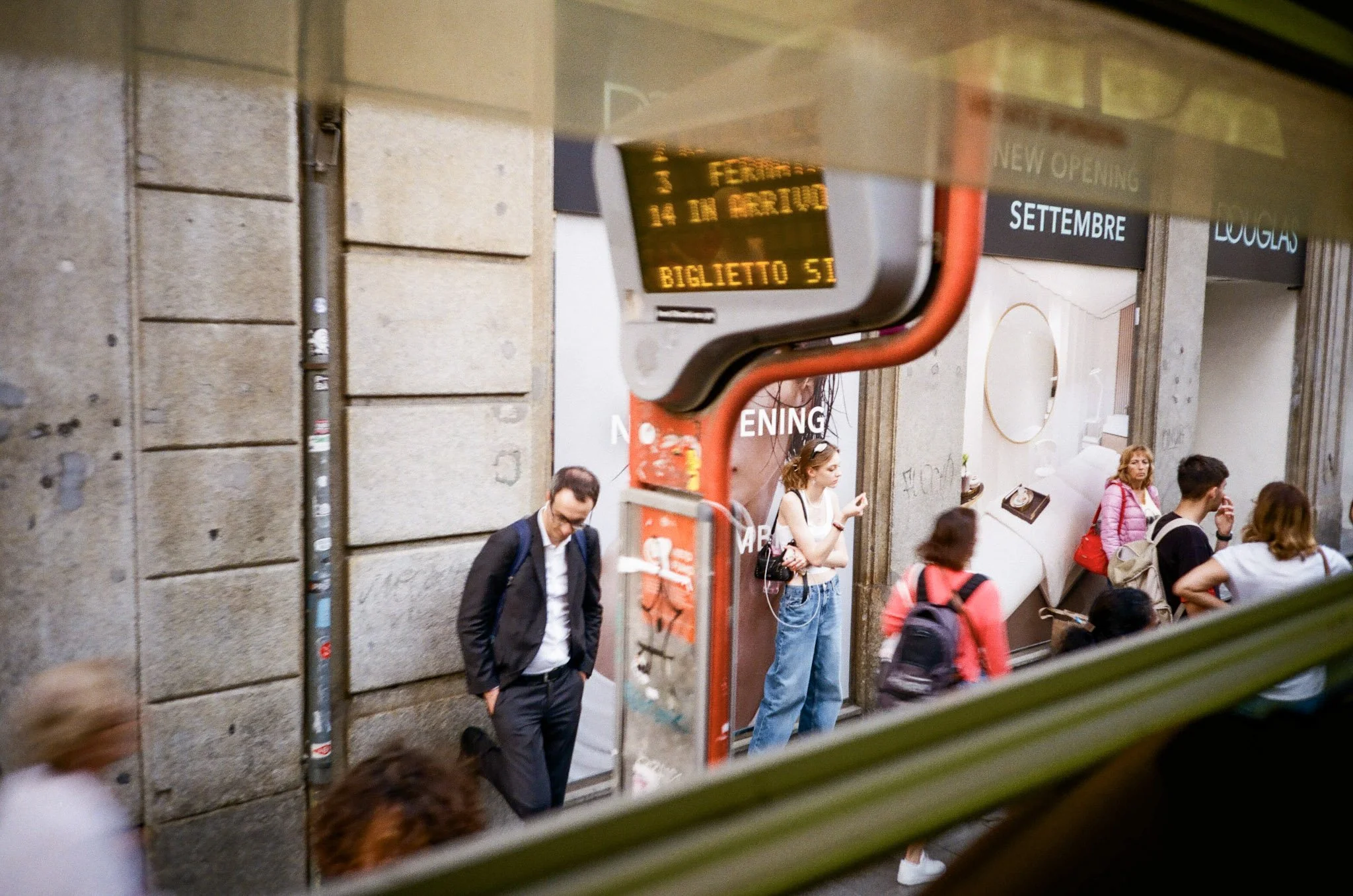 People waiting outside a building, seen through a window with a sign and a digital display at the top, in an urban setting.