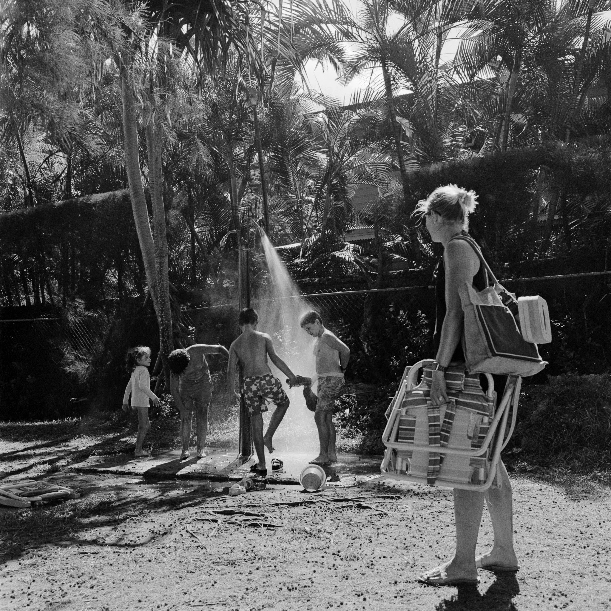 A woman with a tote bag carrying a striped beach chair and a cooler standing on the sand, watching children playing under a shower of water surrounded by tropical trees.
