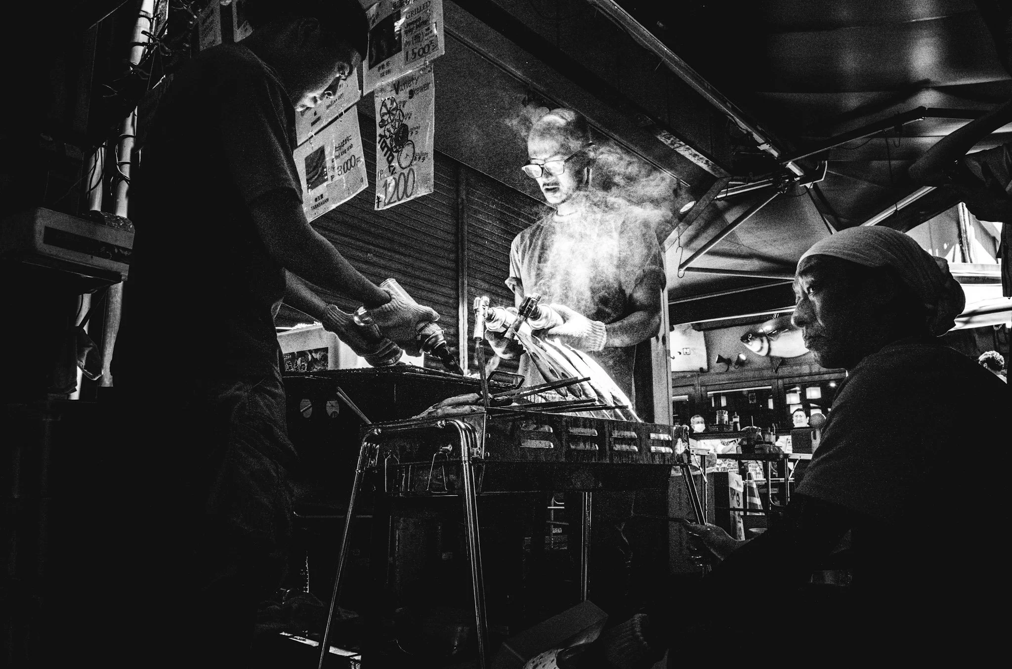 Three people in a street food stall, one grilling meat over a barbecue, another holding bottles, and a woman sitting nearby, all in black and white.