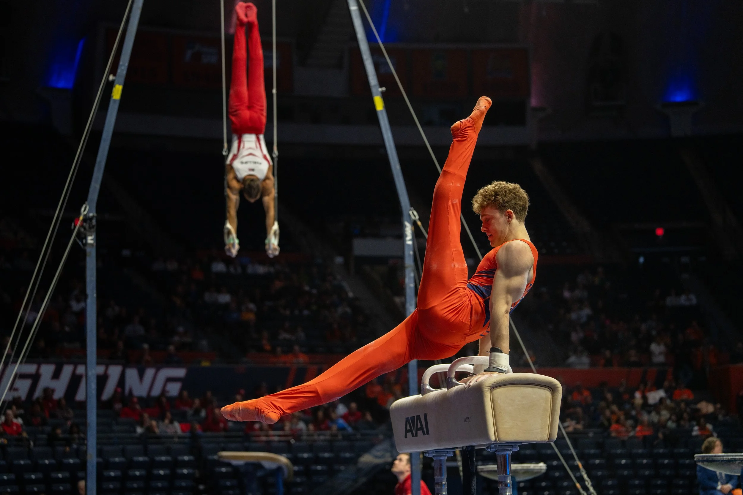 A male gymnast in an orange uniform performing a routine on the pommel horse, with another gymnast in red and white hanging upside down in the background at a gymnastics competition.