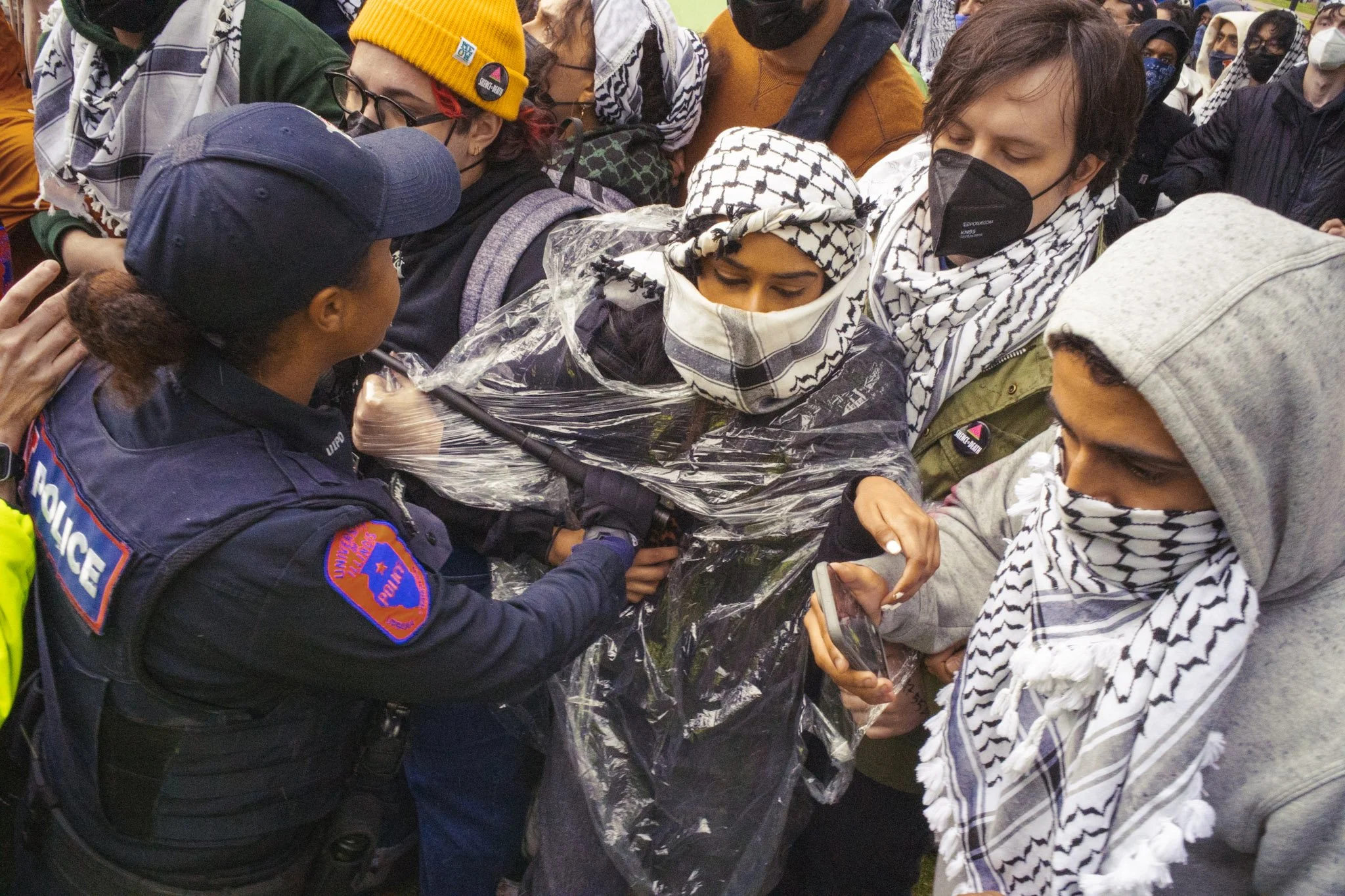 A group of people wearing Palestinian scarves and face masks face off with a police officer. The police officer is holding a microphone and appears to be in the middle of speaking or trying to control the crowd. The scene looks tense, with the crowd 