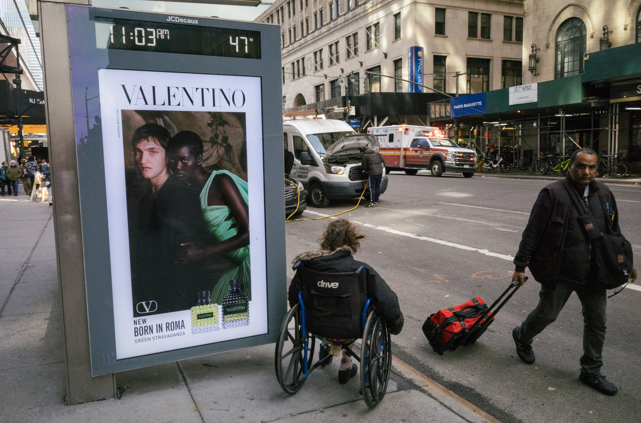 Street scene with people walking, including a woman in a wheelchair and a man pulling a suitcase, on a city sidewalk. There's a digital billboard advertising Valentino perfume and a parked emergency vehicle with flashing lights. Buildings and shop si