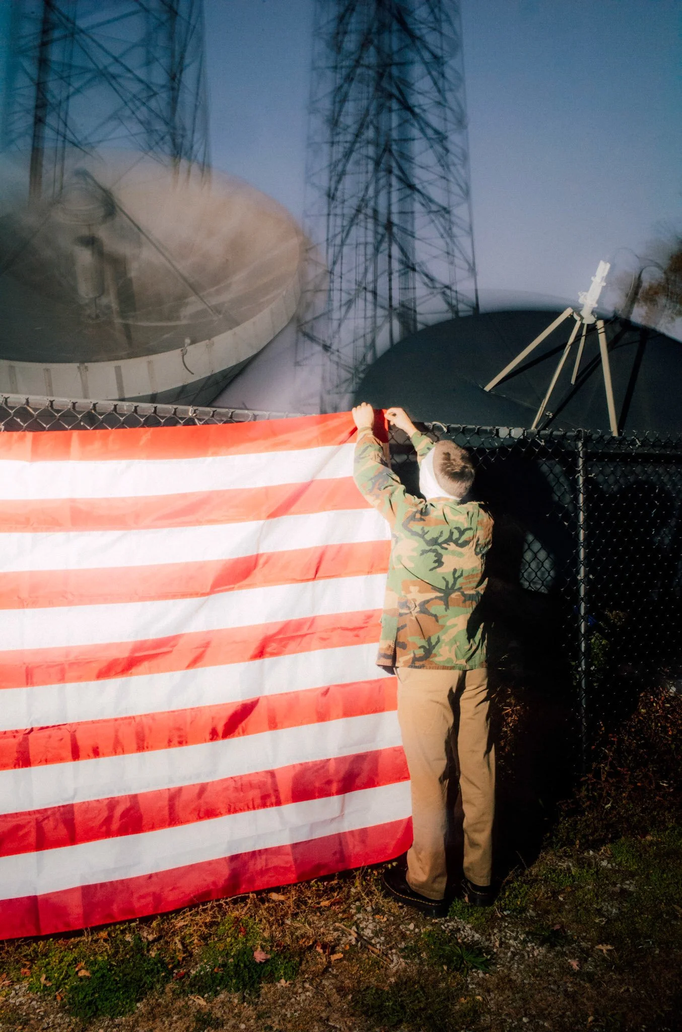 A person in camouflage clothing and tan pants hangs an American flag on a black chain-link fence near radio towers and large satellite dishes during dusk.