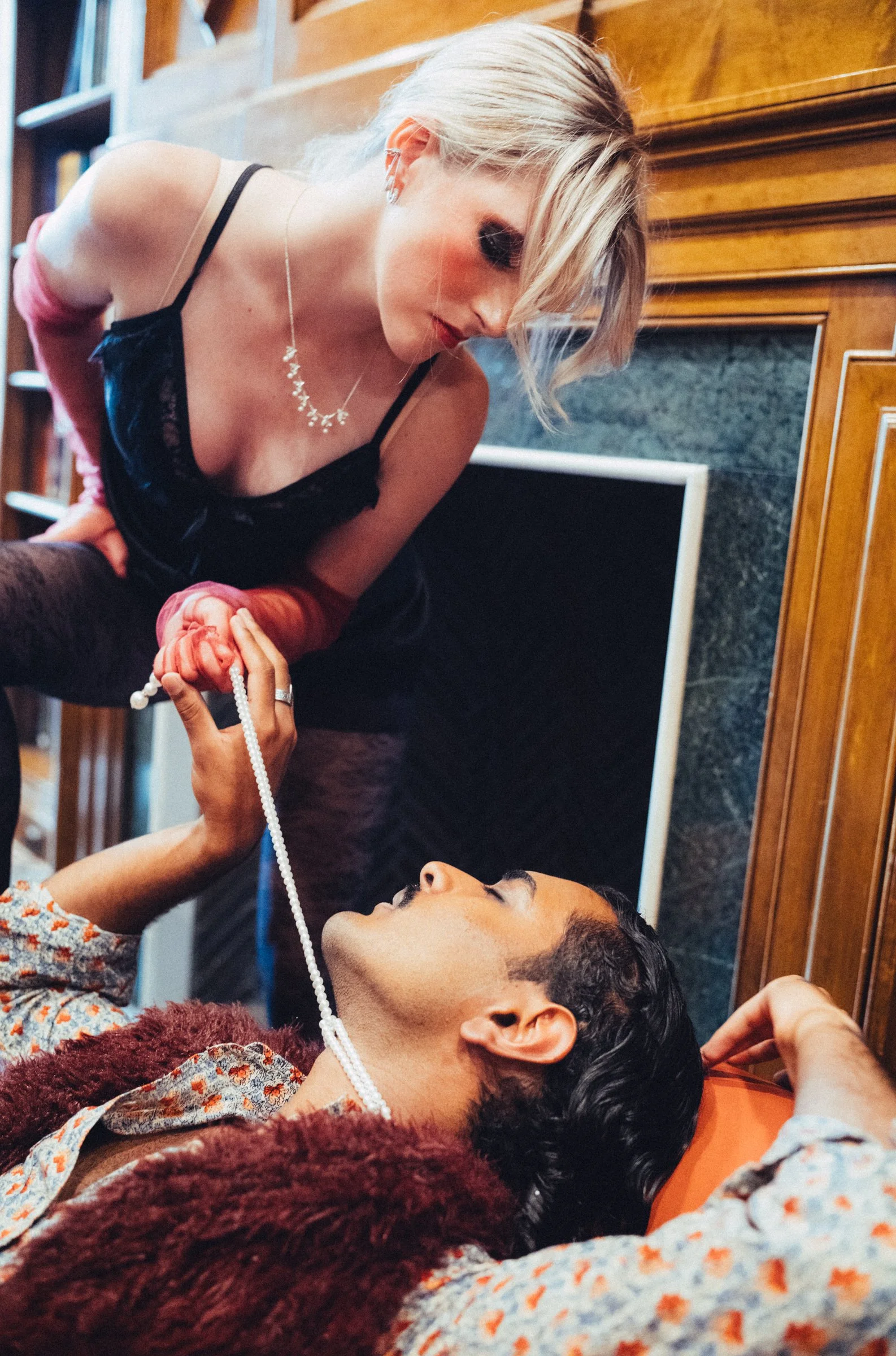 A woman with blonde hair wearing black and pink clothing is leaning over a man lying on the floor, holding a pearl necklace. They are indoors with wooden walls and a bookshelf in the background.