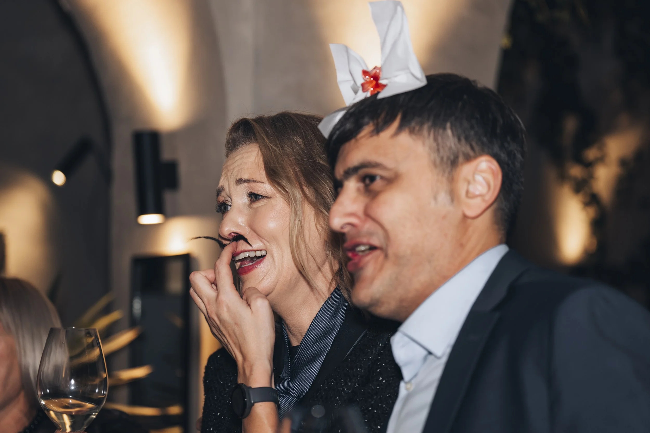 A woman with a fake mustache and a man wearing a party hat at a social gathering.