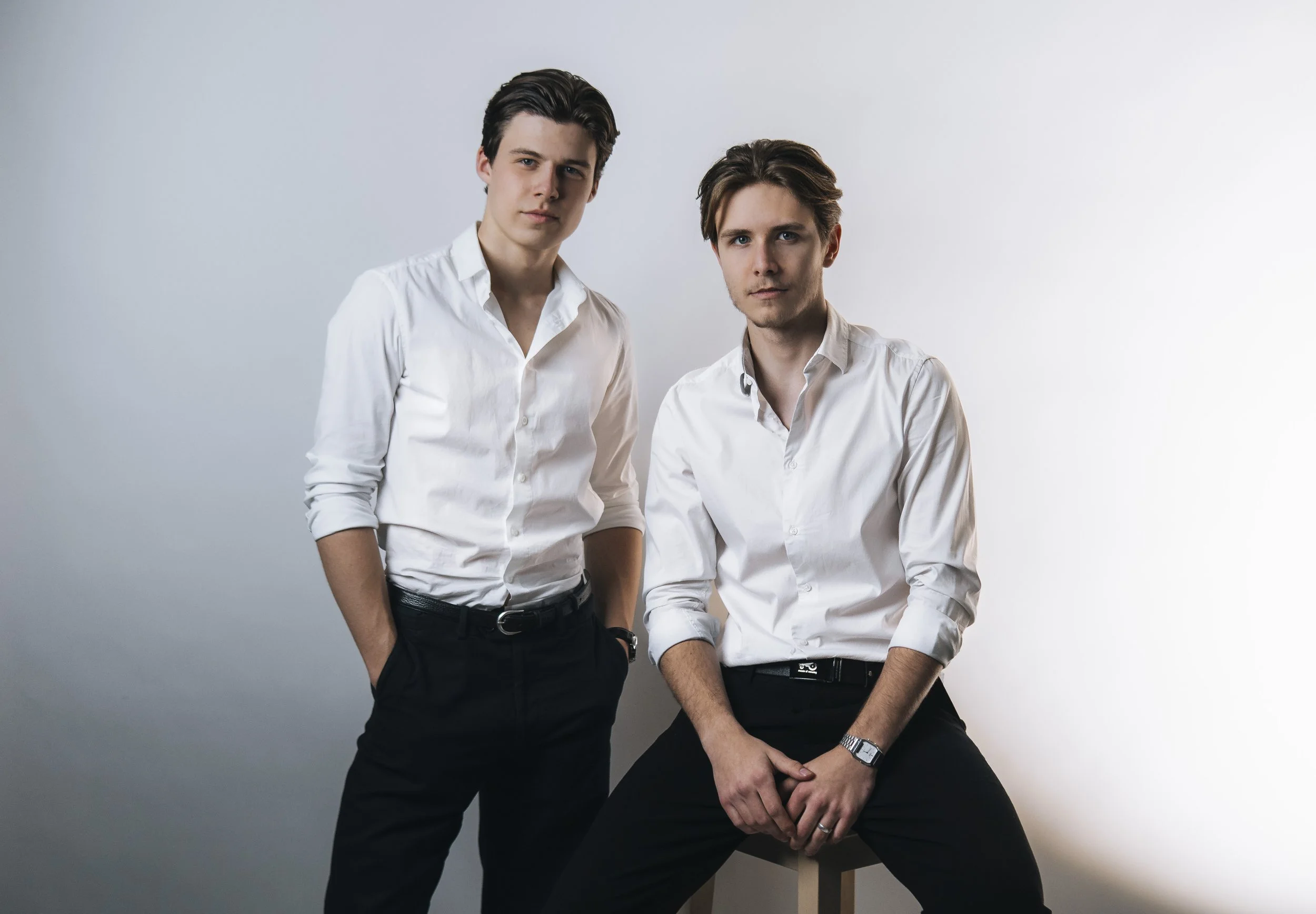 Two young men in white shirts and black pants pose against a plain light-colored background, one standing with hands in pockets and the other sitting on a stool.