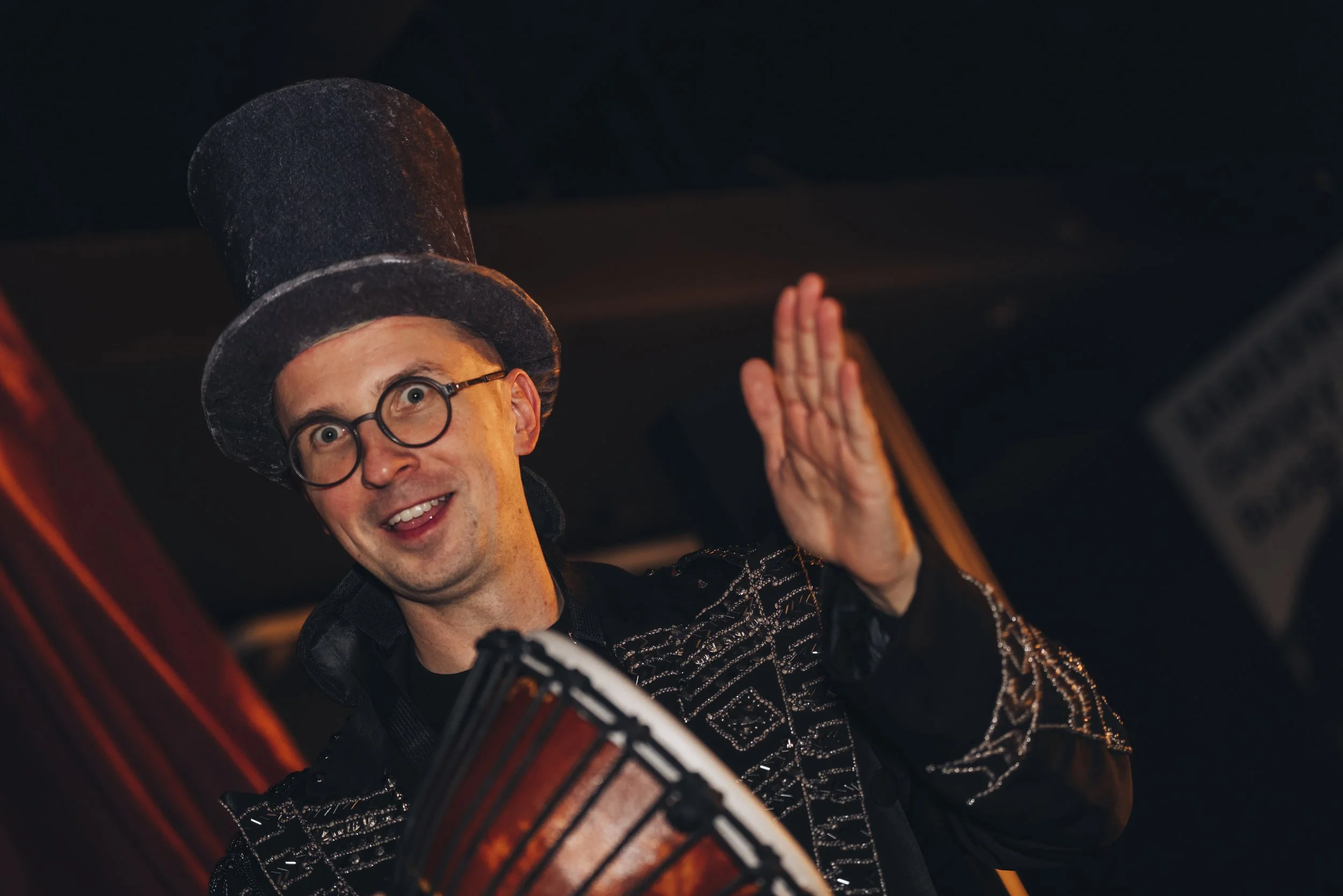 A man wearing a tall black top hat, round glasses, and a decorative black jacket, smiling and waving while playing a drum.