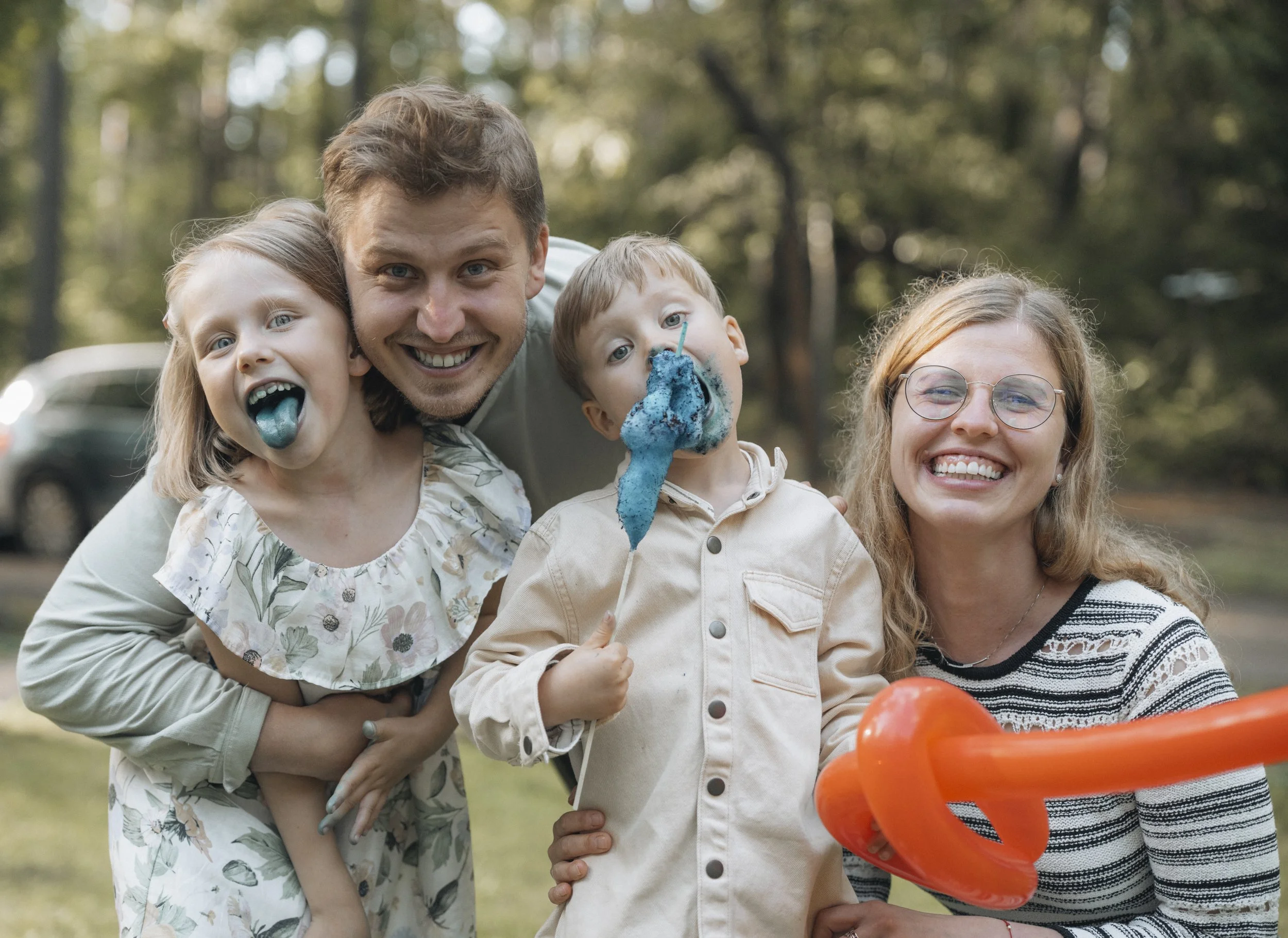 A family of four outdoors smiling and enjoying a celebration. The father and son are covered in blue cake, and the mother is holding orange balloons.