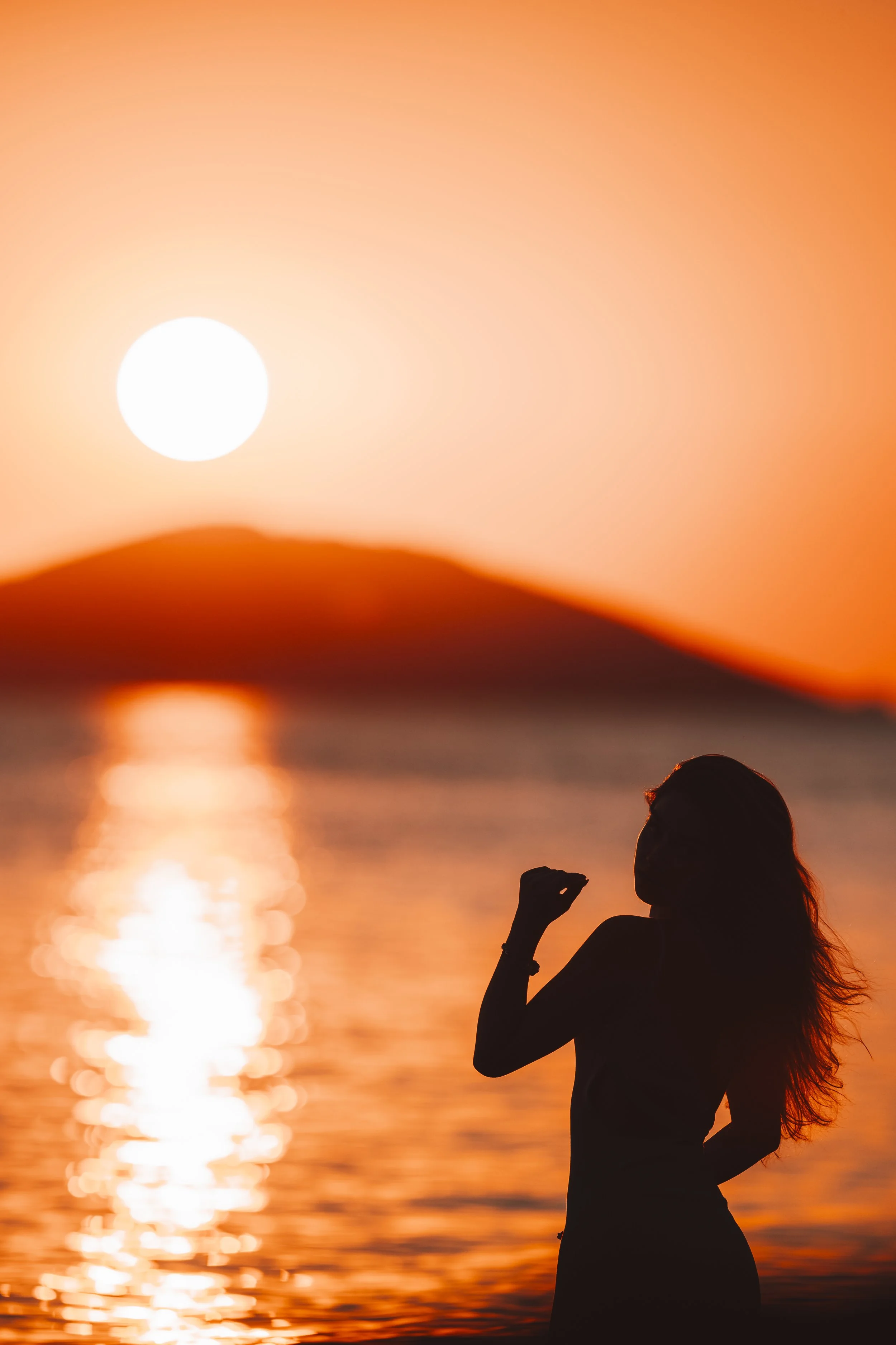 Silhouette of a woman standing in water during sunset, with the sun low in the sky and a mountainous landscape in the background.