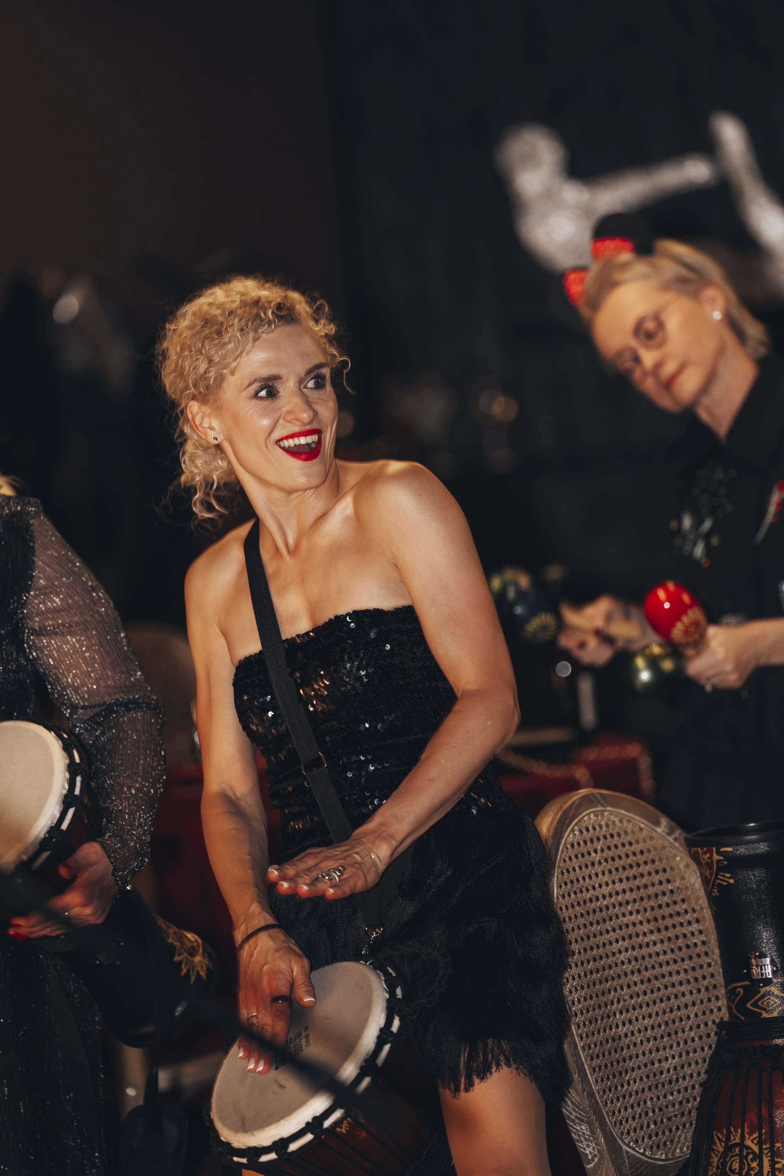 A woman with curly blonde hair in a black strapless sequined dress plays a drum during a festive event, smiling and engaging with others.