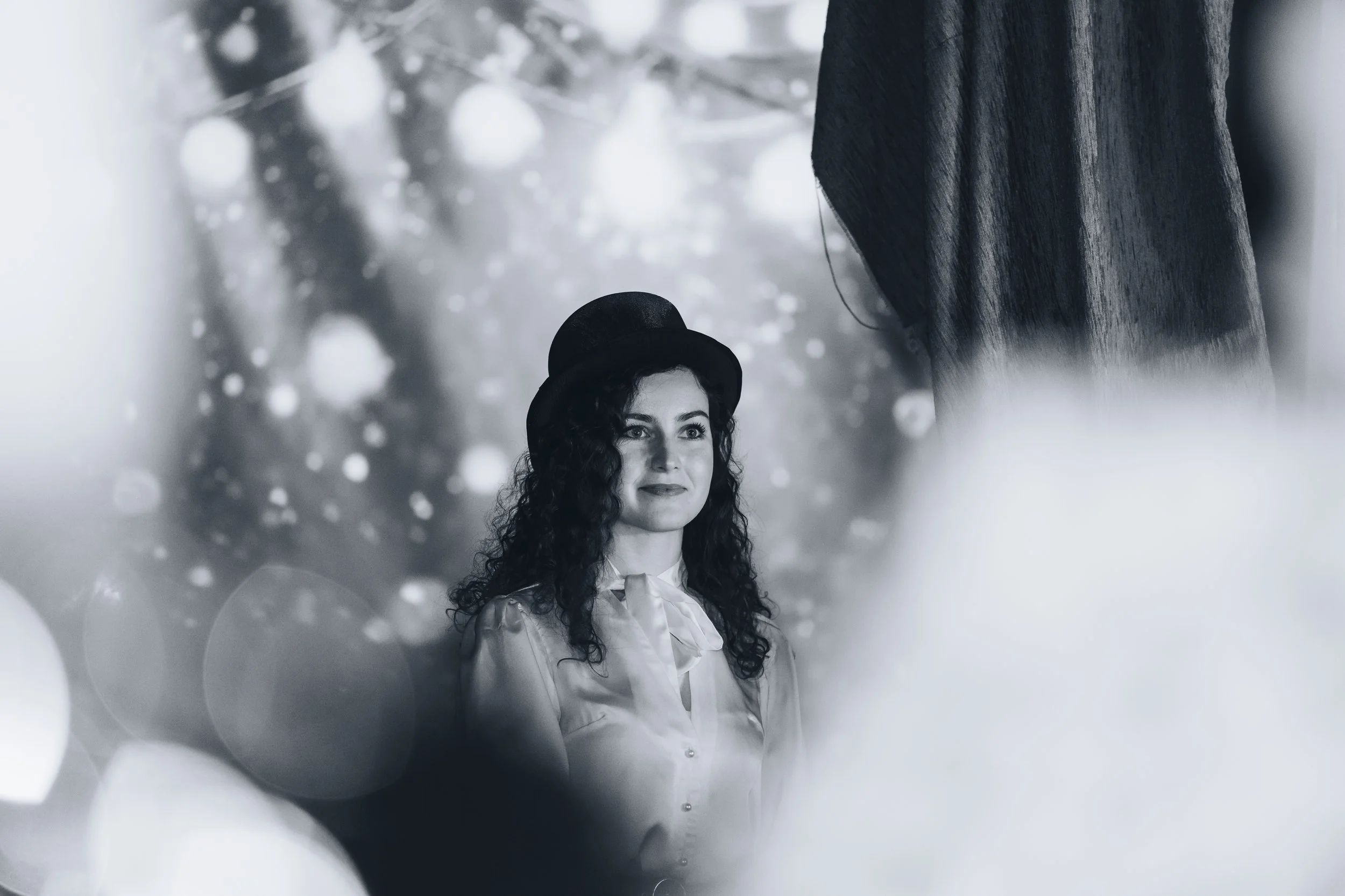 A woman with curly hair wearing a hat and a blouse with a bow tie, looking at a person who is mostly out of the frame. The background has a bokeh effect with circular light spots.