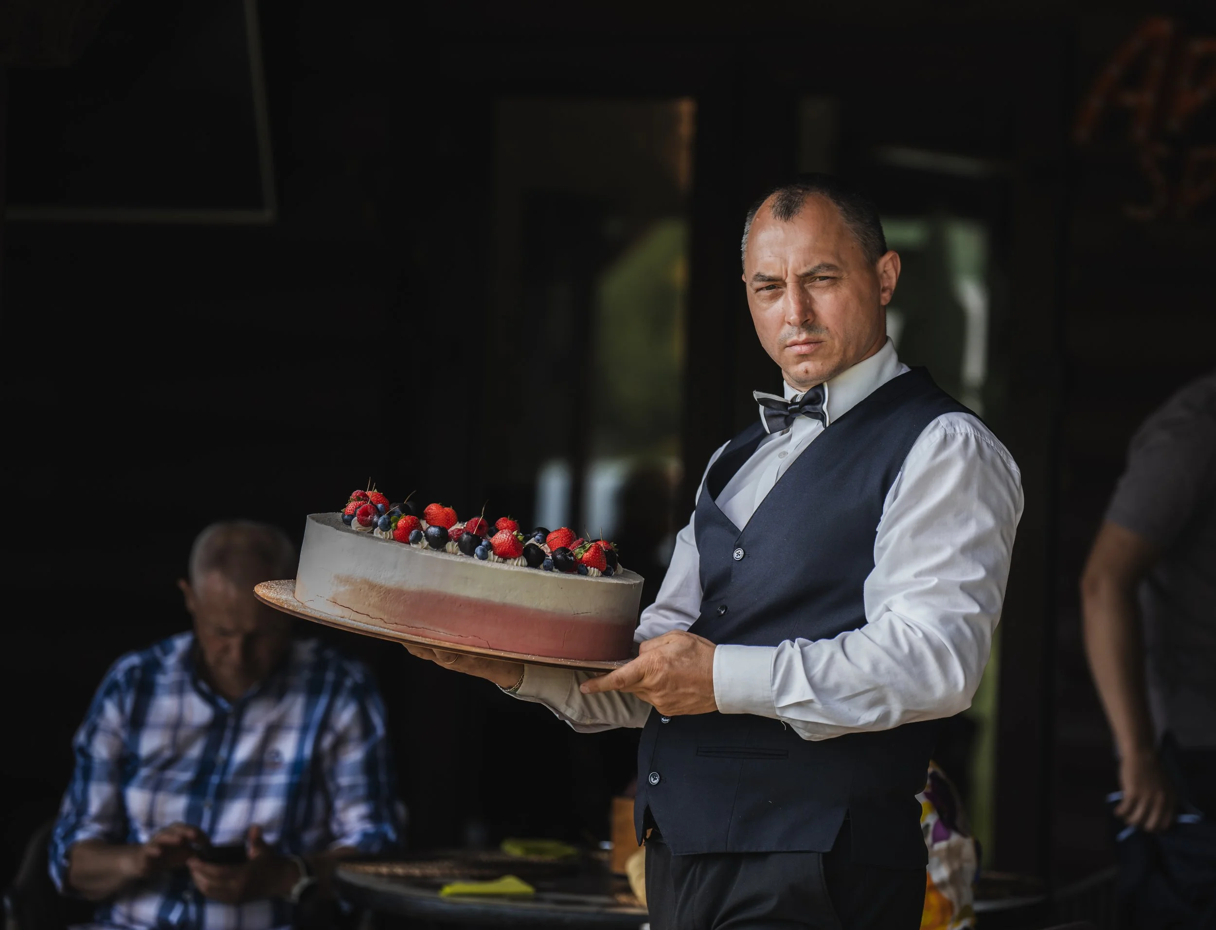 A man in formal attire, including a vest and bow tie, holding a large cake decorated with berries at what seems to be a celebration or event.