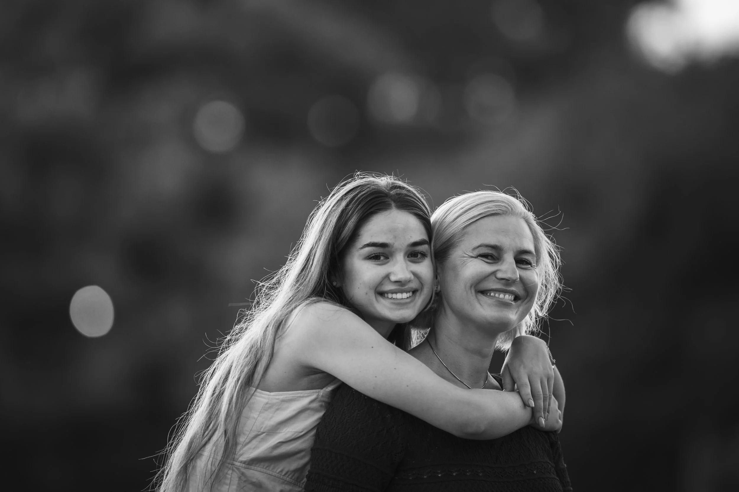 Black and white photo of a young girl hugging an older woman, both smiling warmly at the camera, outdoors with blurred background.