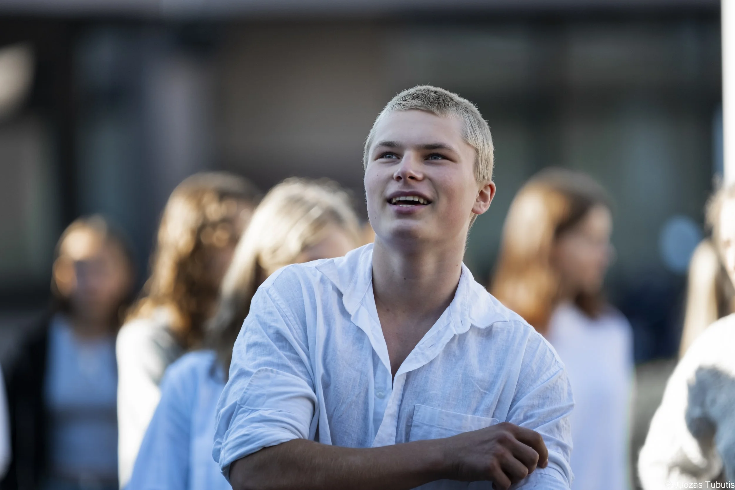 A young man with short blonde hair and pale skin, wearing a white shirt, smiling and looking slightly to the side. Several blurred people are in the background.
