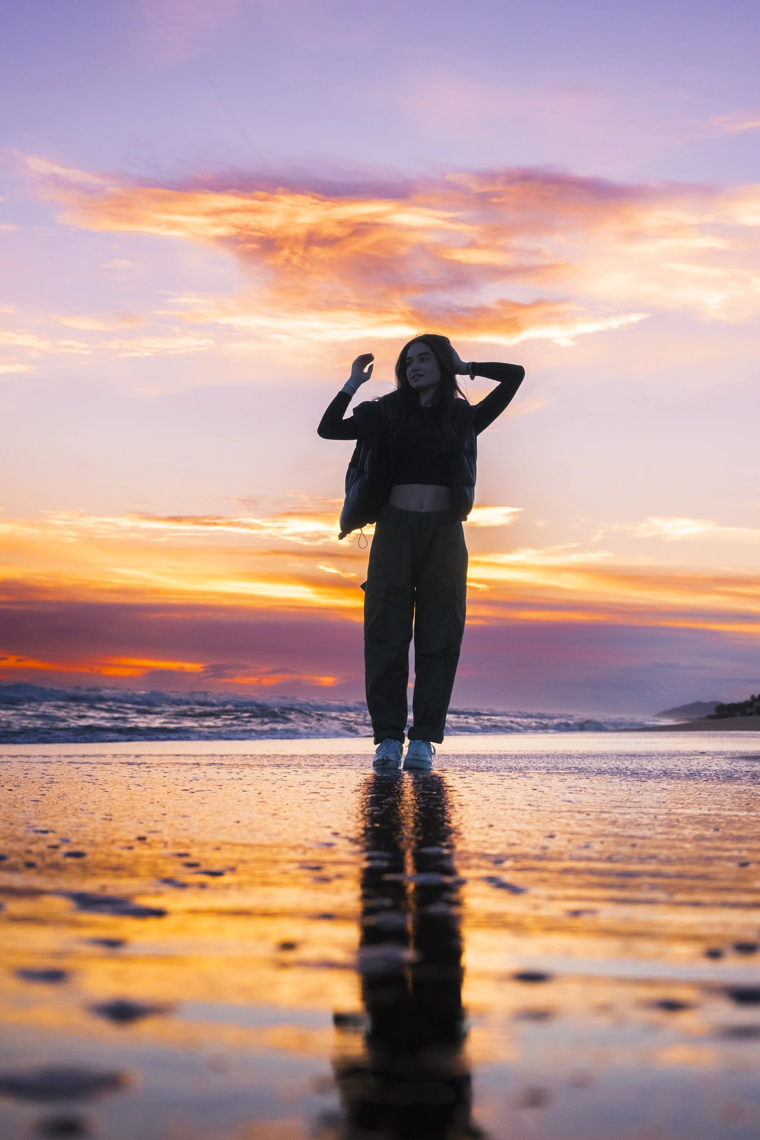 A woman stands on a reflective wet beach at sunset, with colorful sky and clouds in the background.