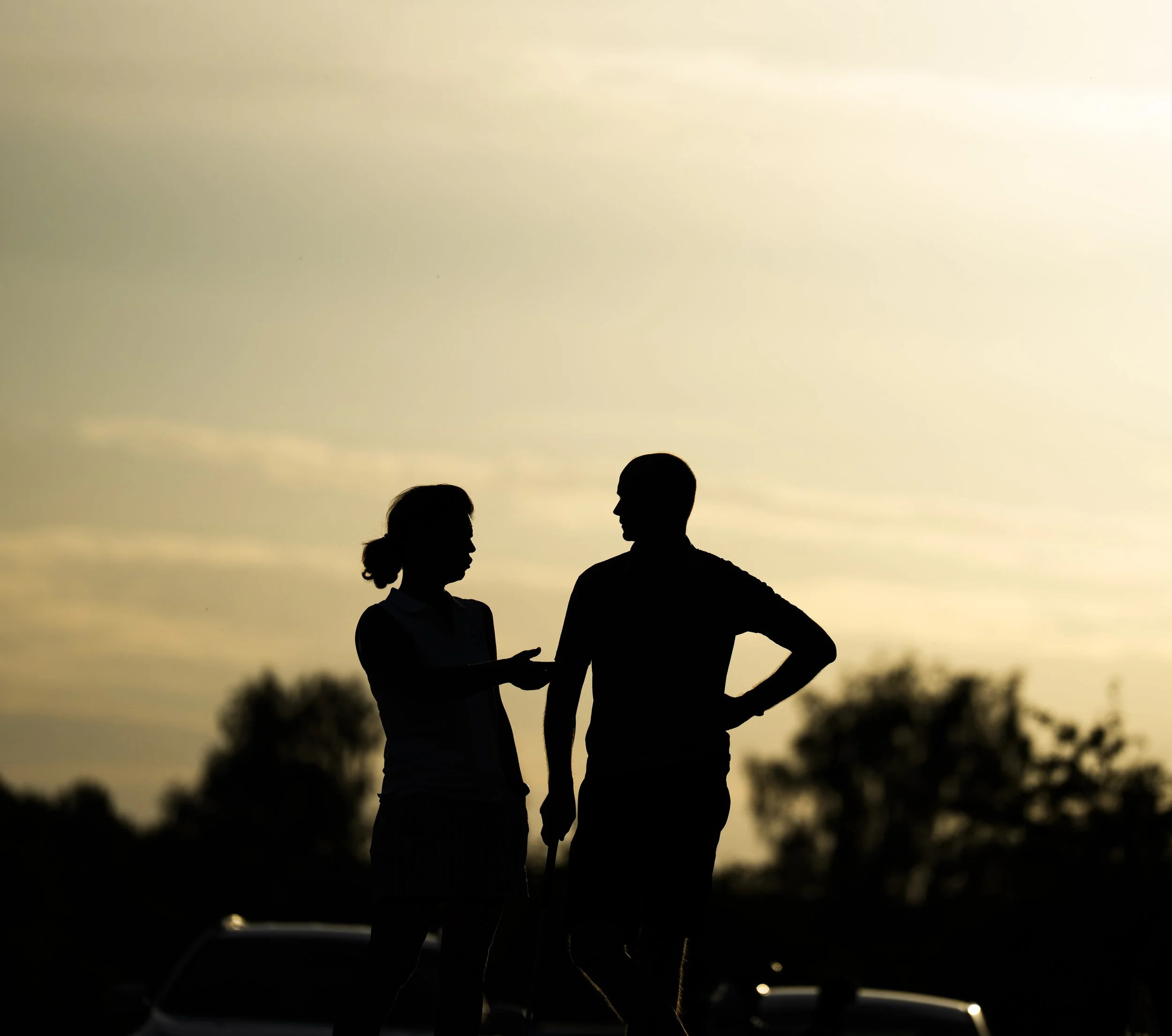 Silhouette of a man and woman having a conversation outdoors during sunset, with trees and cars in the background.