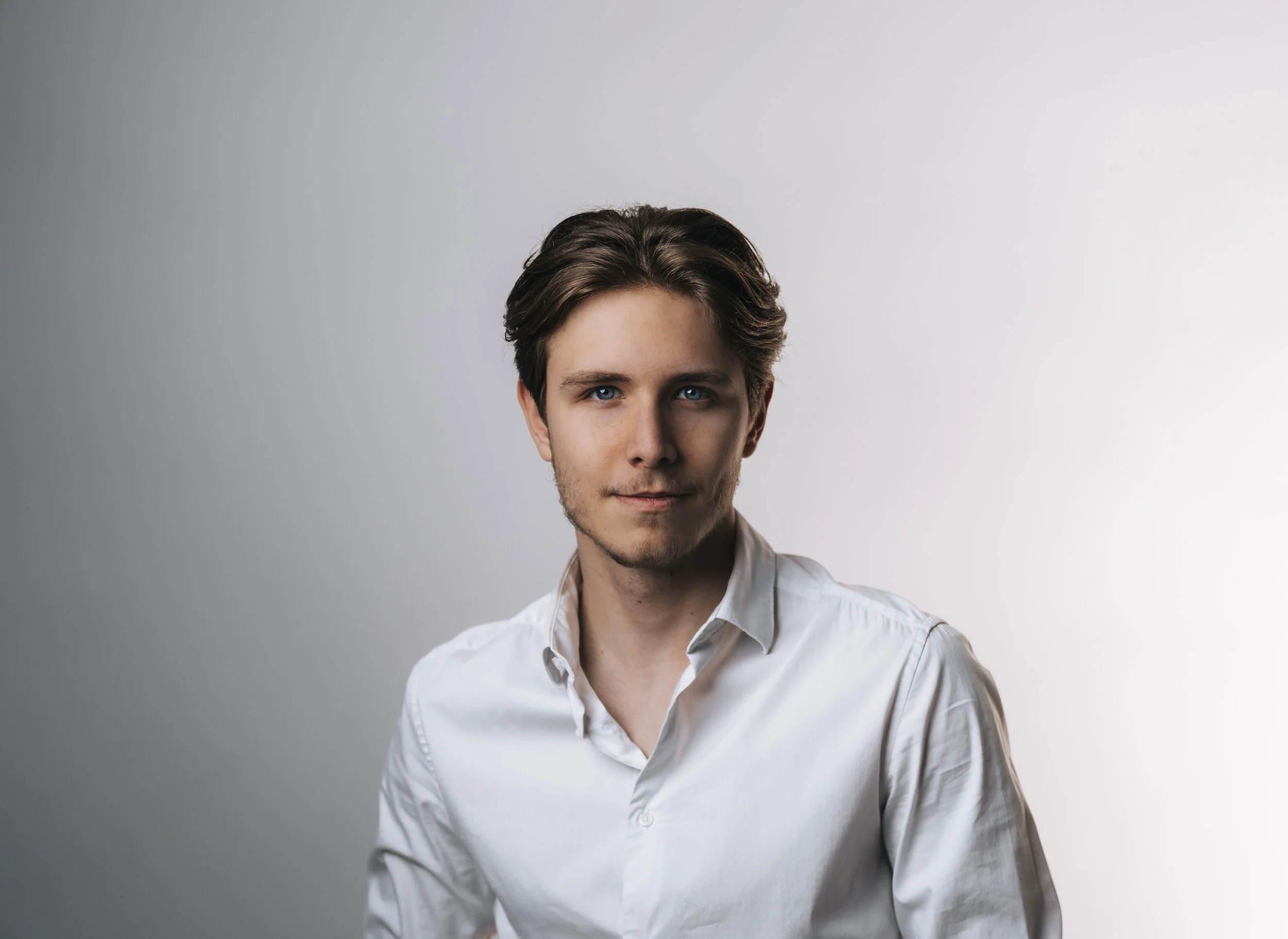 Young man with brown hair and blue eyes wearing a white shirt, looking at the camera against a plain gradient background.