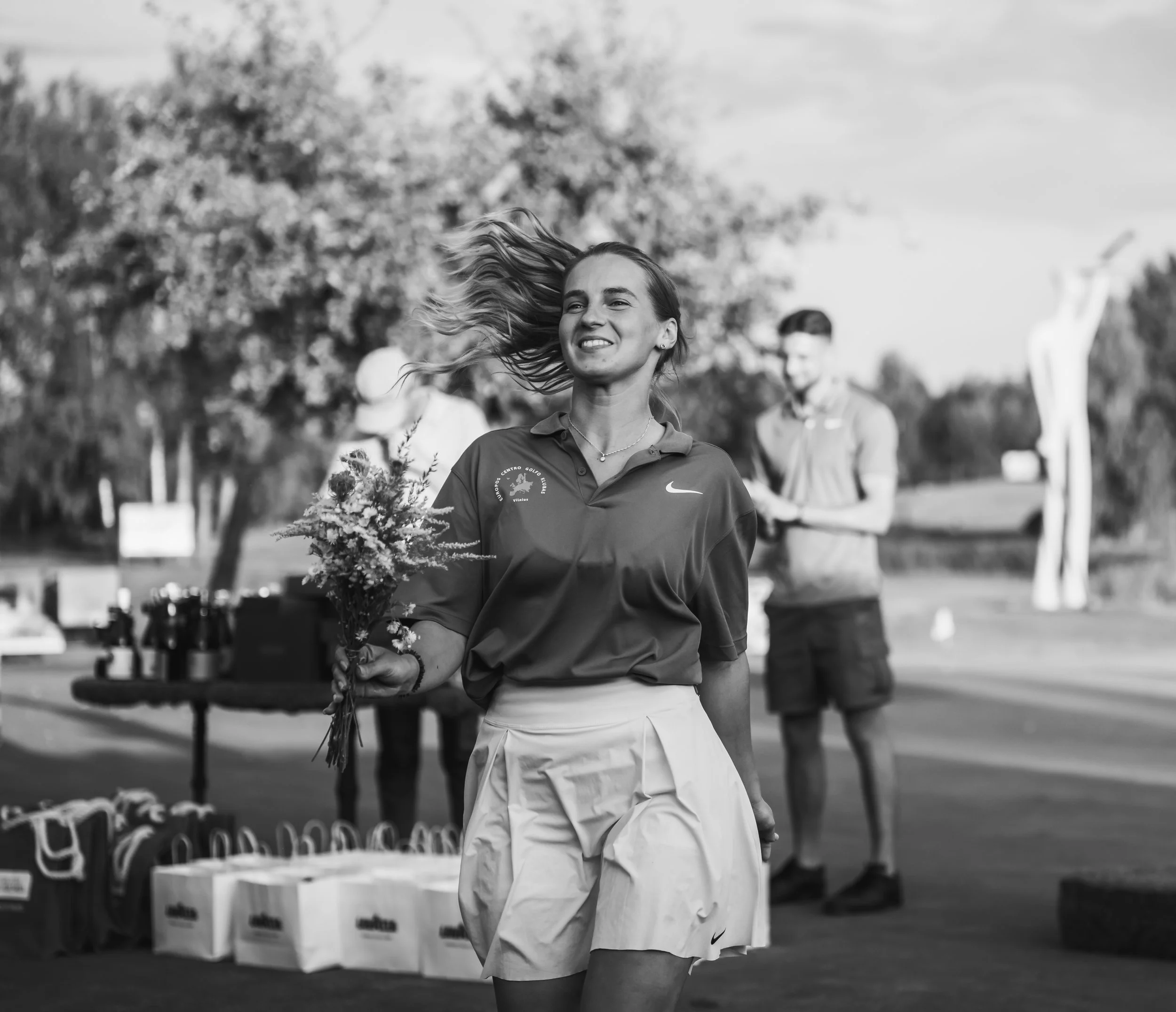 A young woman smiling and holding a bouquet of flowers outdoors, with two men and trees in the background during daytime.