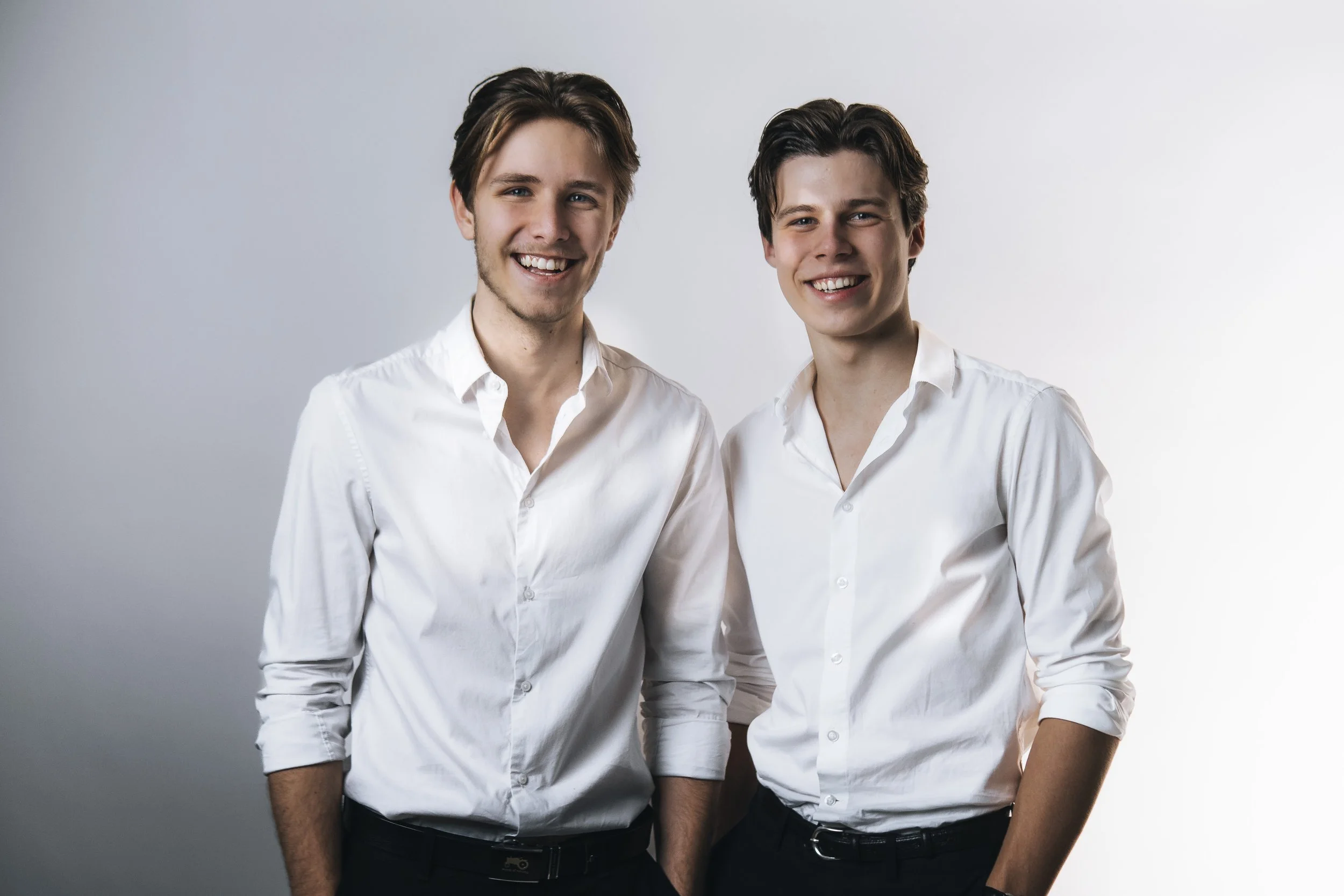 Two young men with similar appearance standing side by side, smiling at the camera, wearing white shirts against a neutral background.