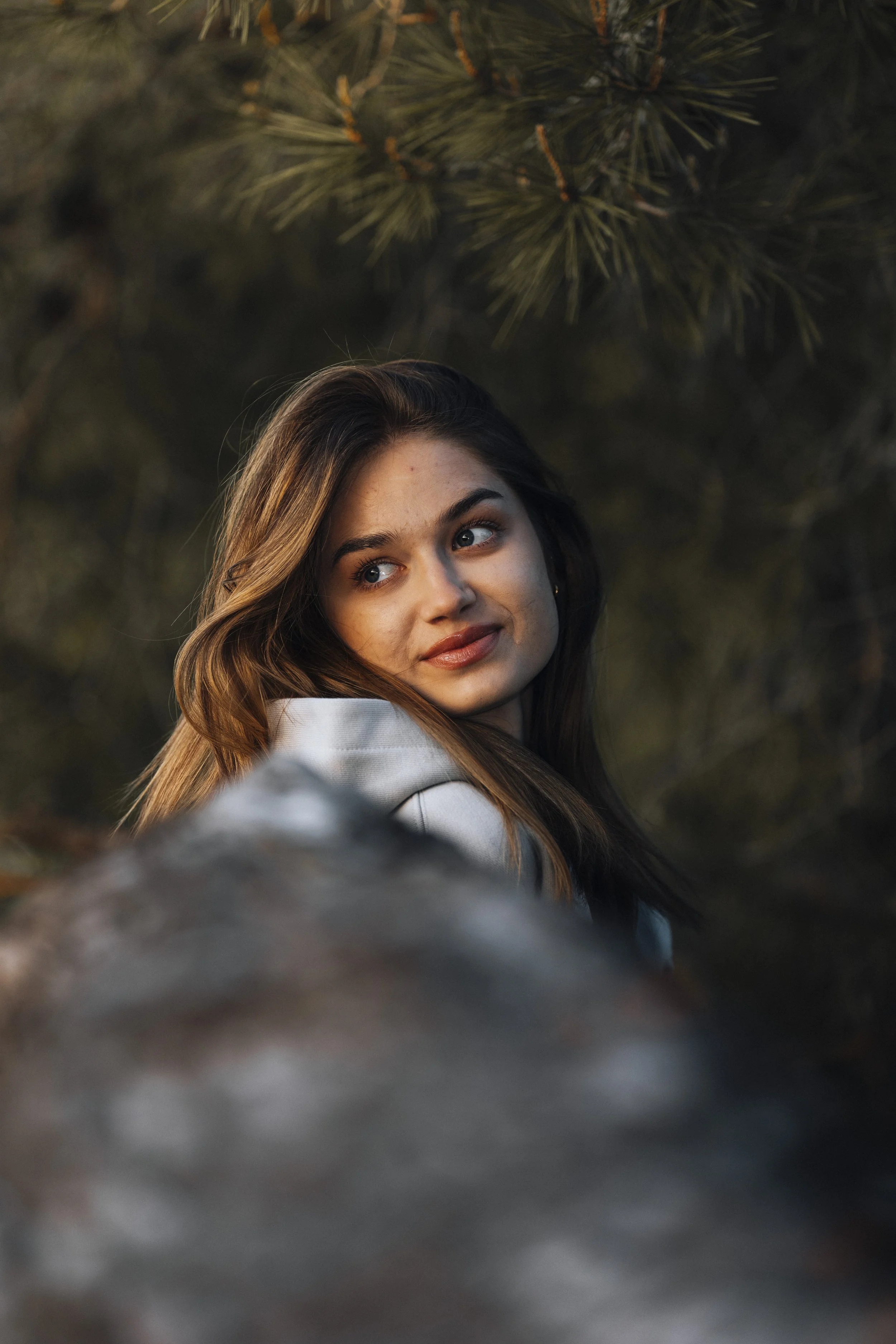 A young woman with long brown hair and blue eyes looking over her shoulder outdoors near pine trees.