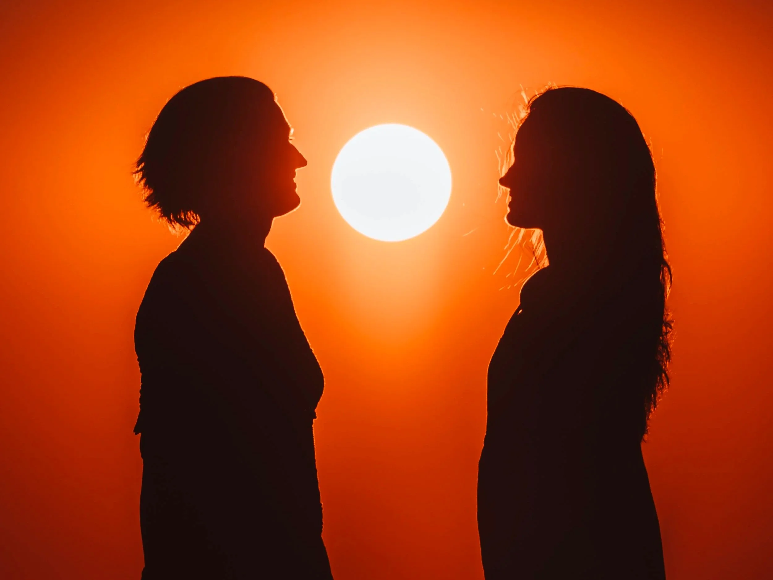 Silhouette of two women facing each other with a bright sun in the background during sunset or sunrise.