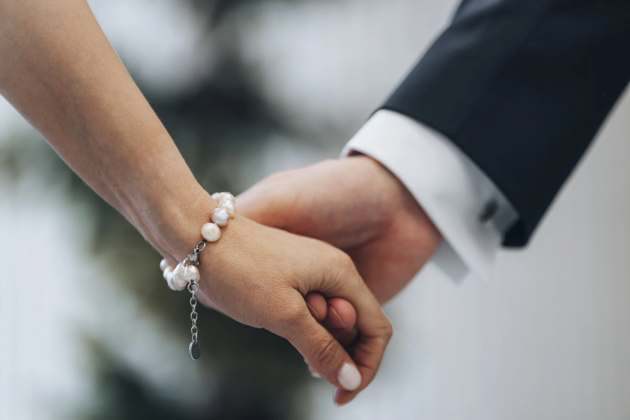 Close-up of a bride and groom holding hands during a wedding, with the bride wearing a pearl bracelet and the groom in a black suit with a white shirt.