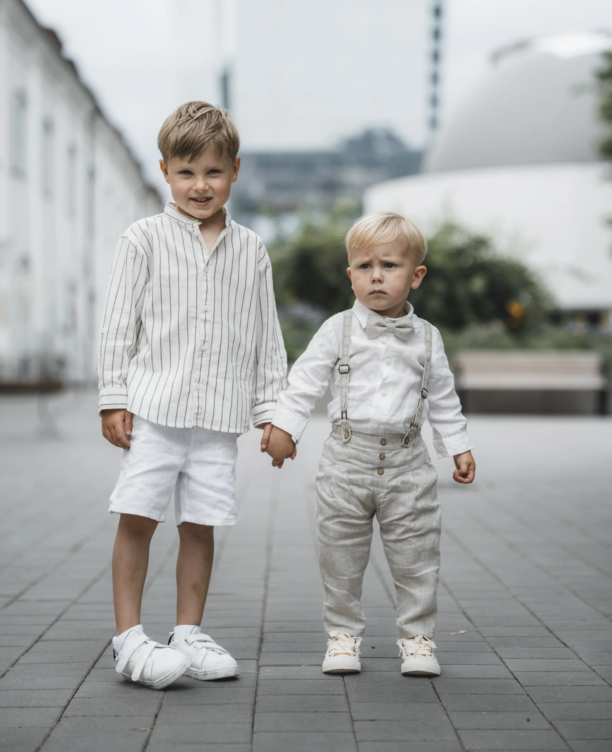 Two young boys holding hands outdoors in a city setting, dressed in light-colored casual and semi-formal clothing with sneakers.