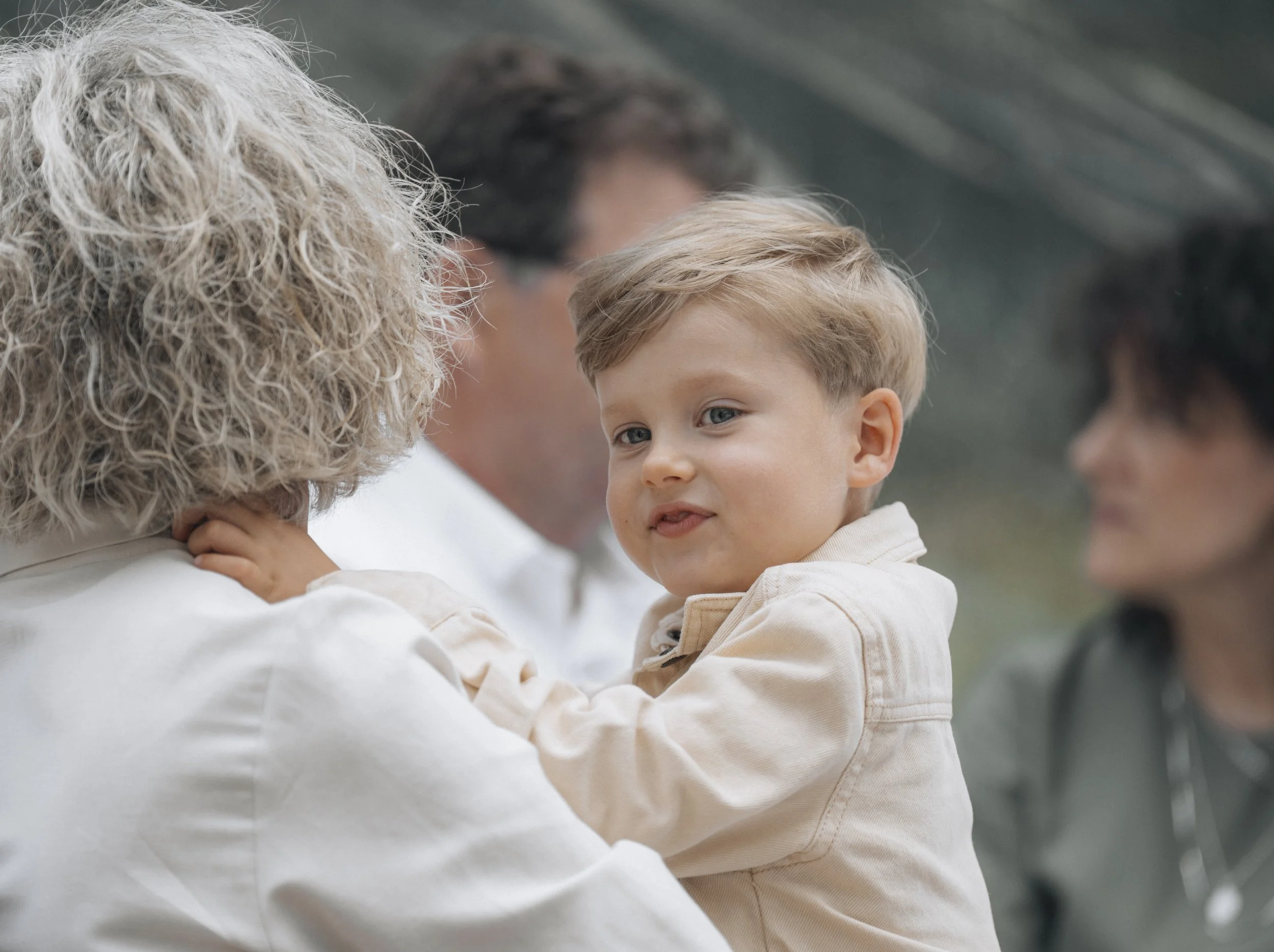 Young boy with light brown hair and blue eyes being held and kissed by an older woman with curly blonde hair, in an outdoor setting with three other adults in the background.