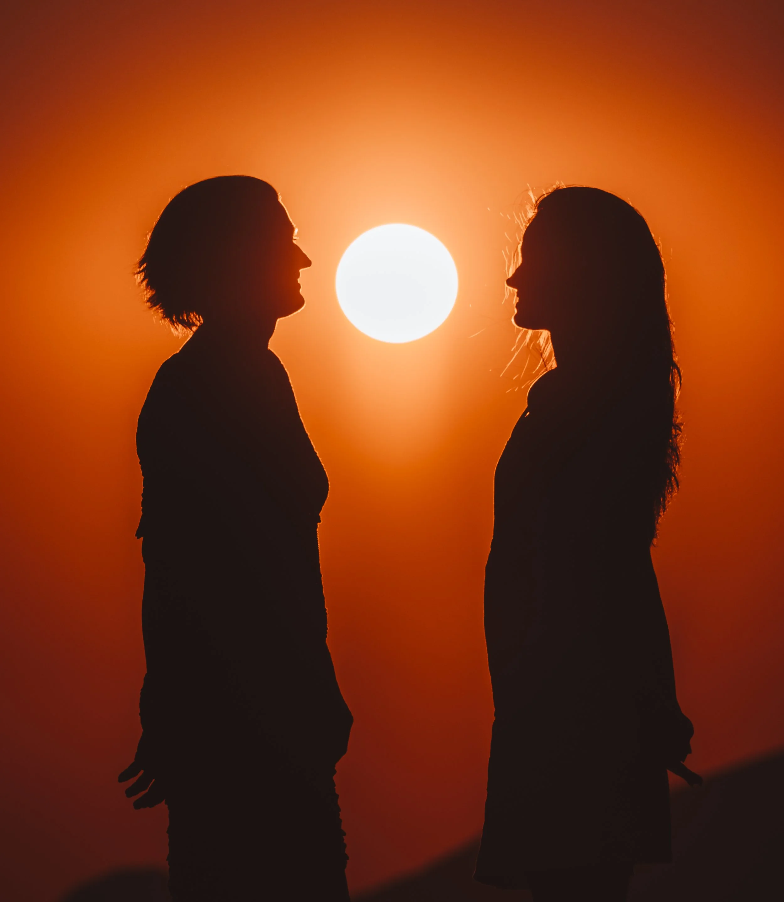 Silhouettes of two women facing each other against a bright setting sun with an orange sky.