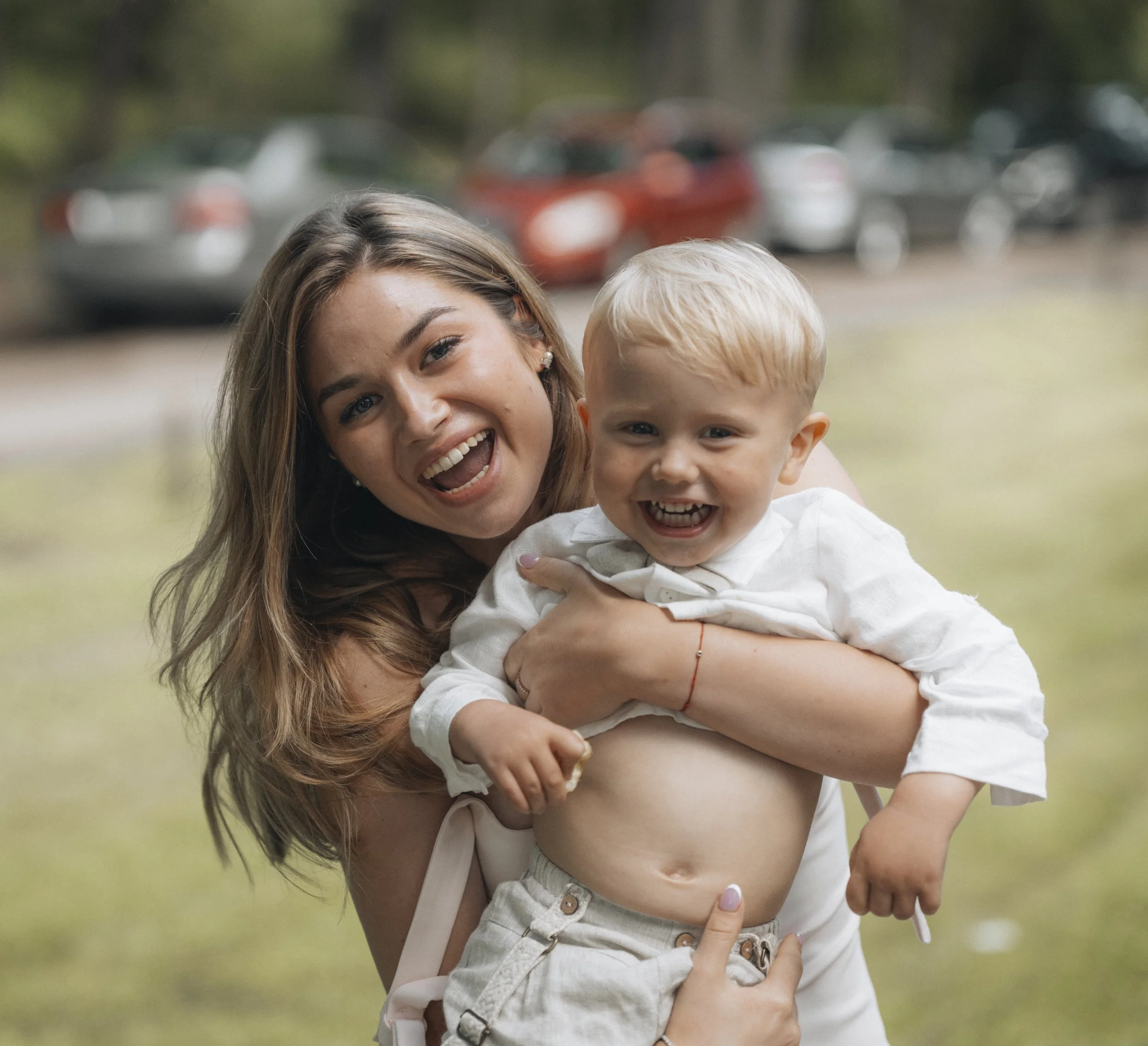 A young woman with long light brown hair is holding a smiling blonde boy in her arms outdoors, with a background of blurred cars and greenery.