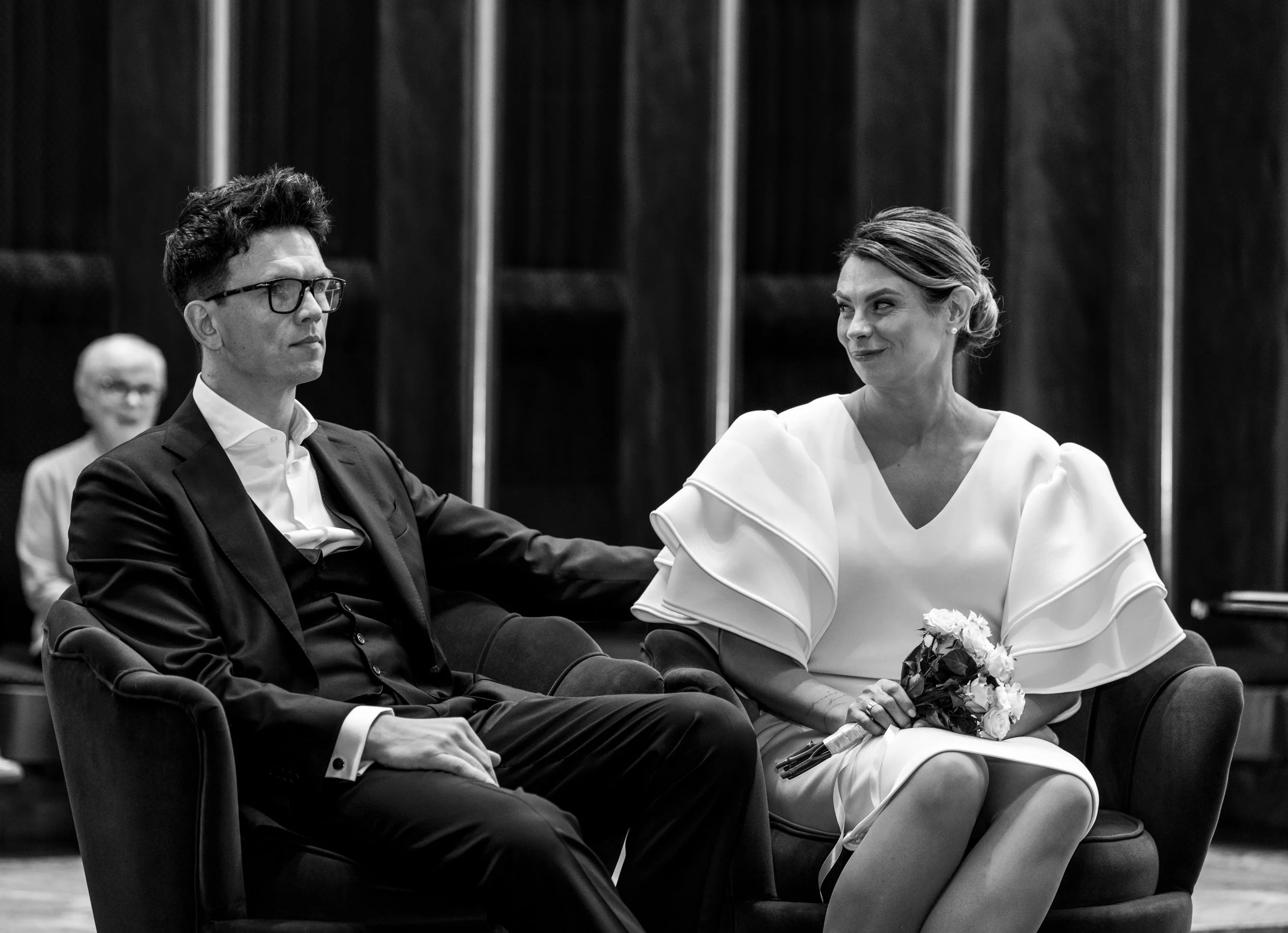 A black and white photo of a man and woman sitting on a sofa, with a woman in the background. The man is wearing a suit and glasses, looking serious. The woman is holding a small bouquet of flowers, smiling faintly, wearing a dress with large, layere