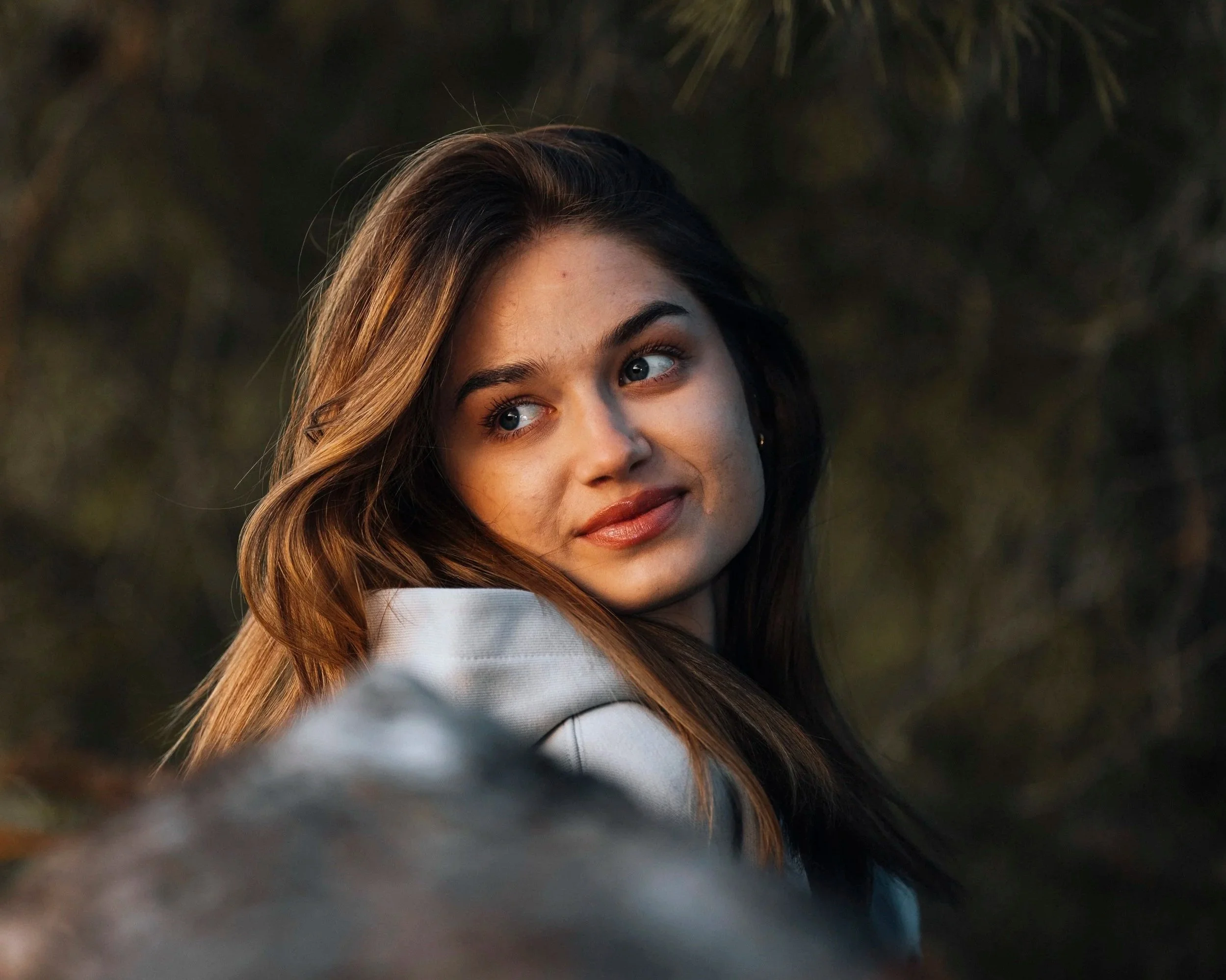 A young woman with brown hair and blue eyes looking over her shoulder, outdoors with blurred greenery in the background, during sunset.