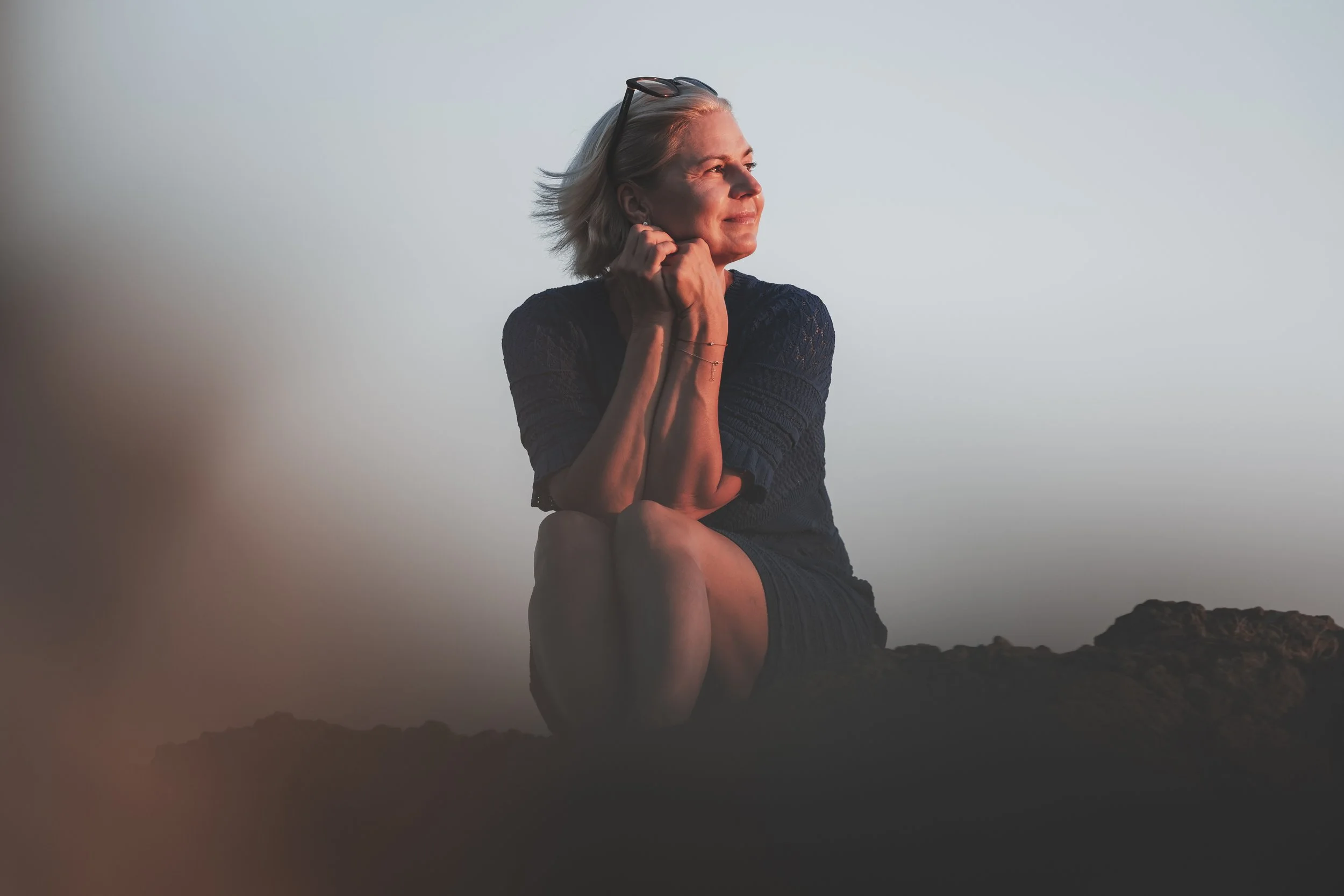 A woman sitting on rocks, gazing into the distance during sunset or sunrise, with sunglasses on her head and a contemplative expression.