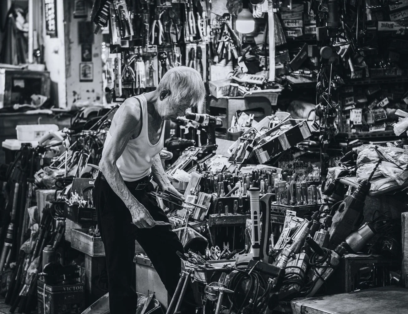 An elderly woman with short gray hair shopping in a cluttered hardware store, examining tools on a table, surrounded by hanging and piled tools and equipment.