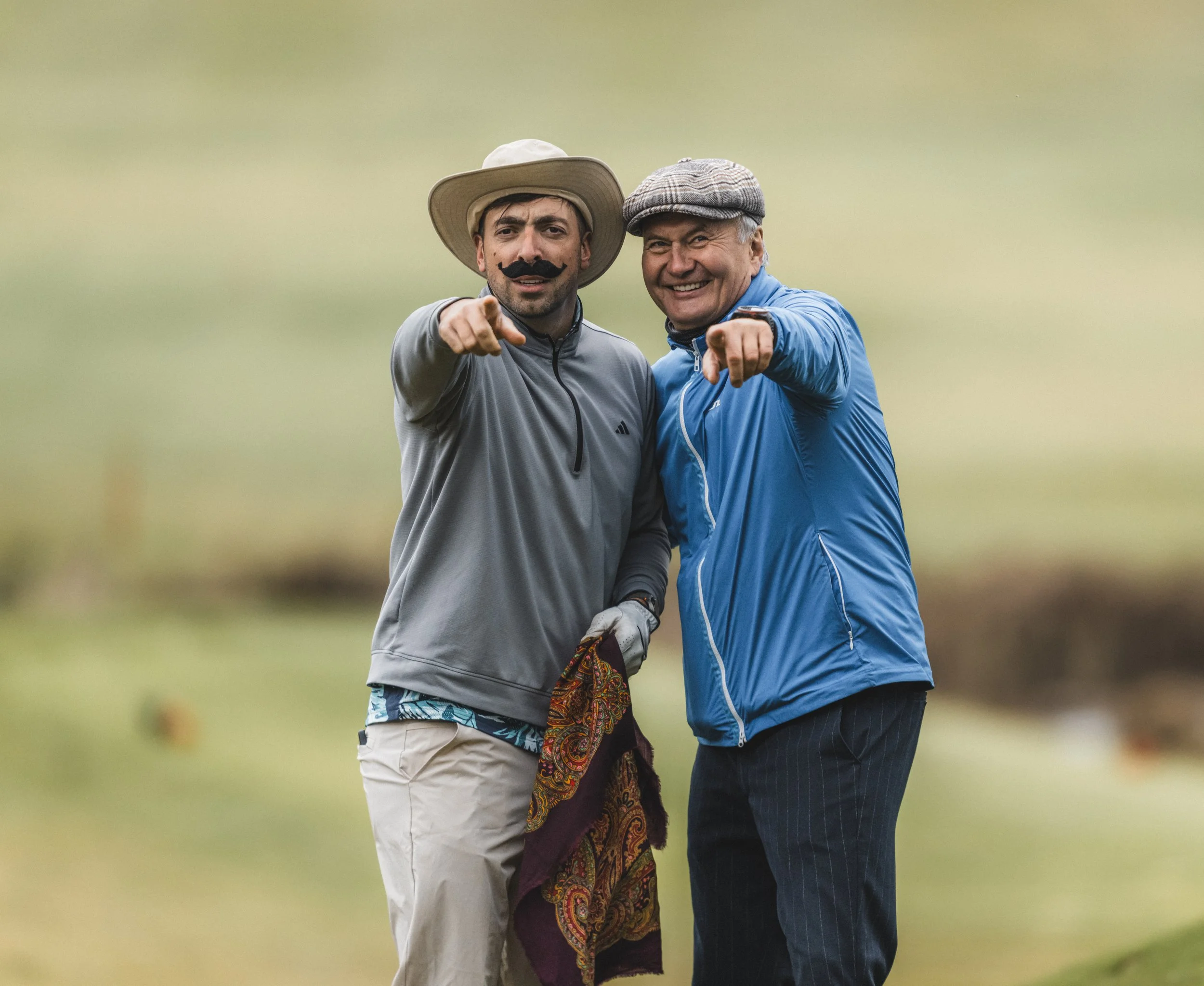 Two men standing outdoors, facing the camera, pointing forward, smiling, on a golf course, with a blurred green background.