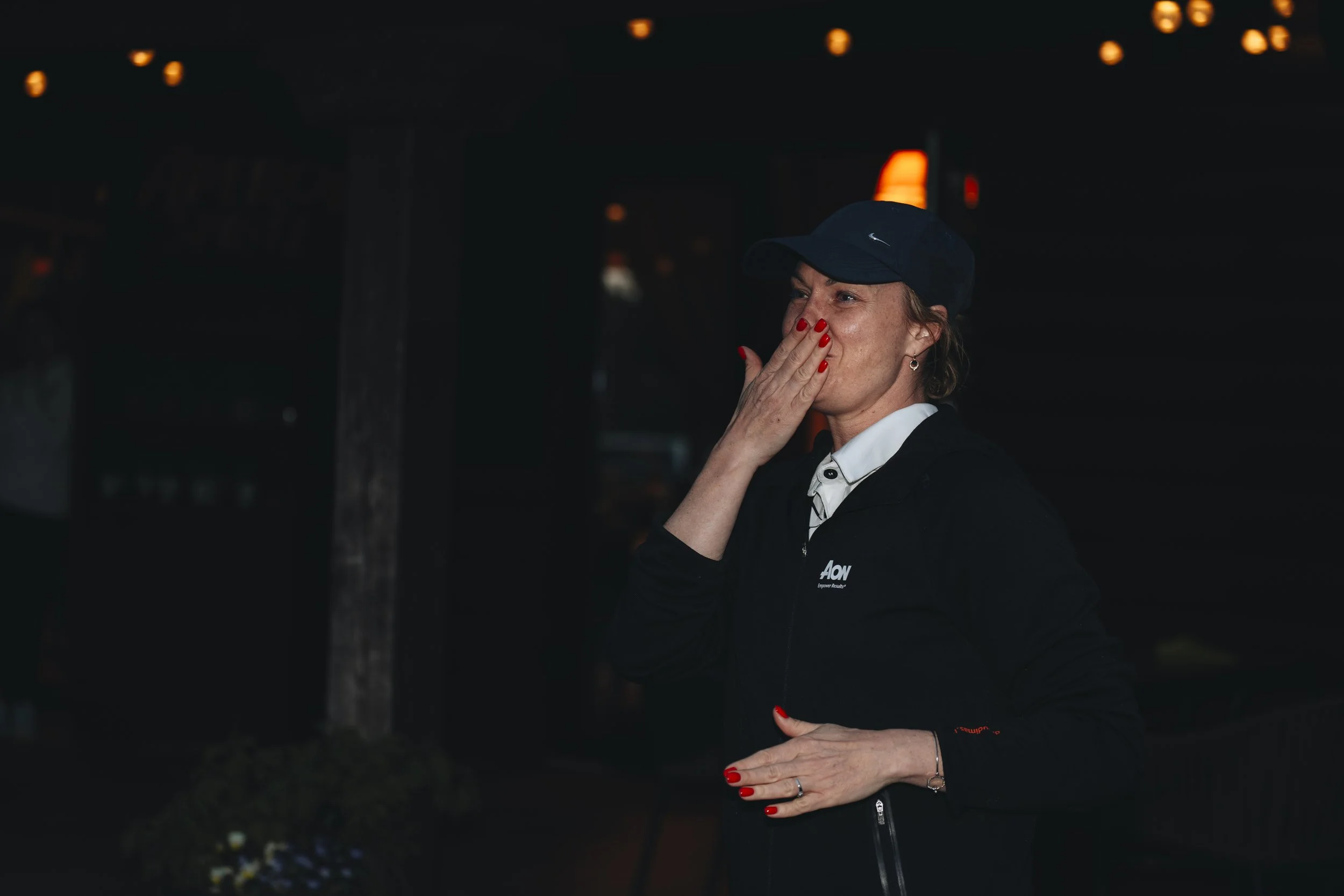 A woman wearing a navy baseball cap and black jacket with an 'AON' logo, holding her hand to her face, smiling, with brightly painted red nails, in an indoor or nighttime setting.