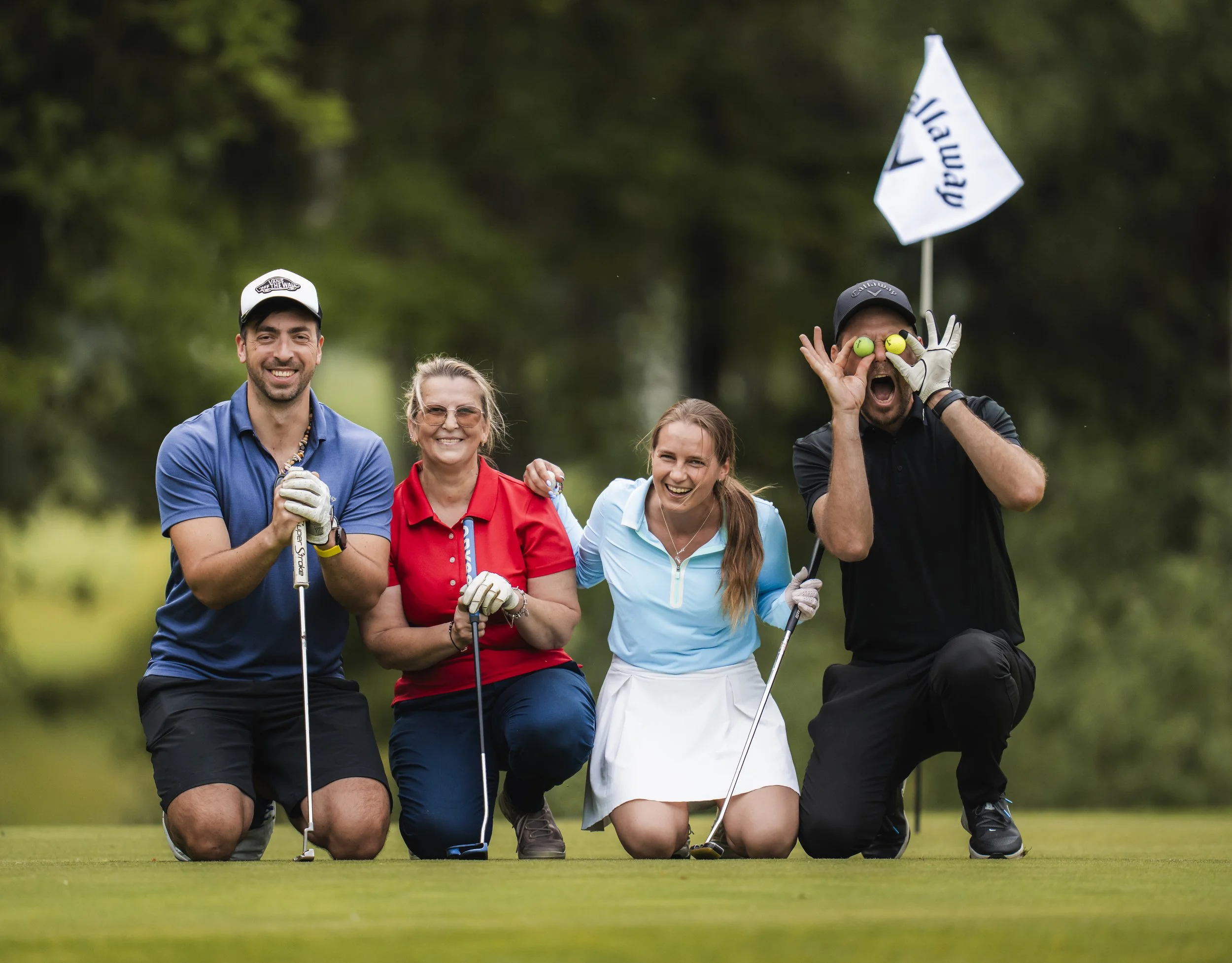 Four people on a golf course smiling and posing for a photo. Two men and two women, dressed in golf attire, with one man holding a flag and the other man holding two golf balls over his eyes.