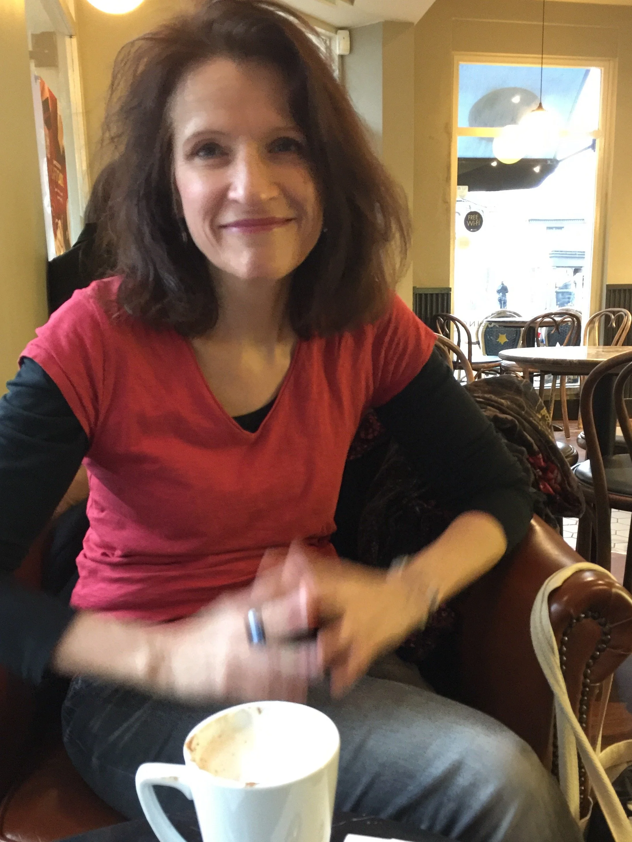 A woman with shoulder-length brown hair, wearing a red t-shirt over a black long sleeve shirt, sitting in a cafe with a coffee mug in front of her, smiling at the camera.