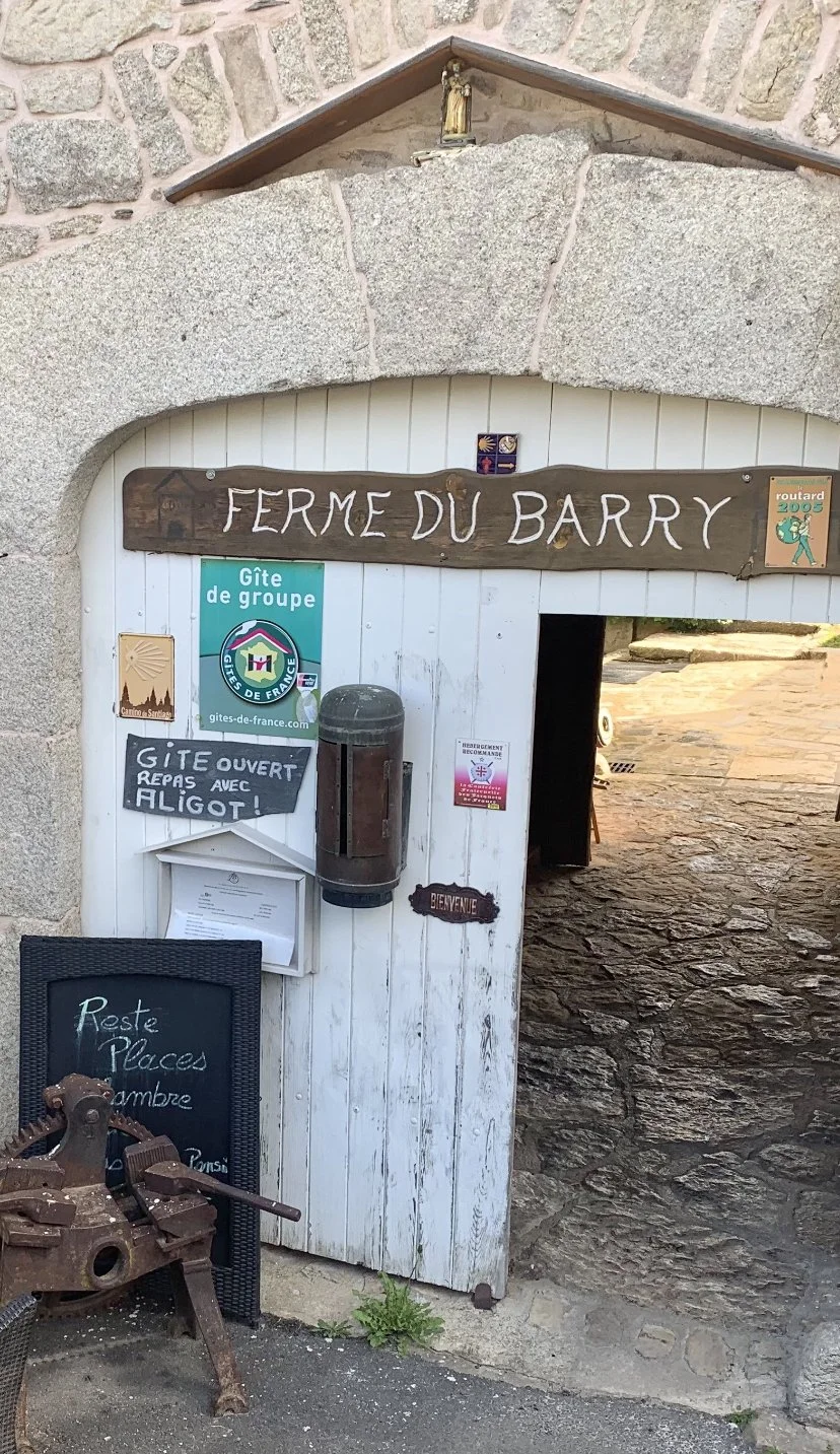 Entrance to Ferme du Barry with signs indicating it is a group campsite open for meals with eel, and decorated with various rustic signs and posters, including a rusty mechanical object at the bottom left.