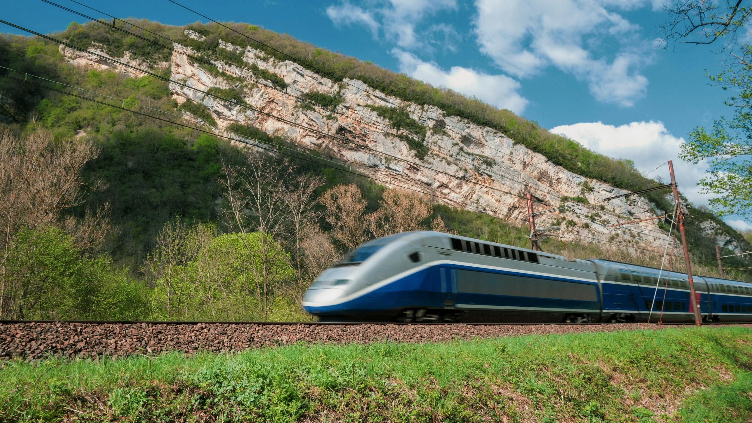 A high-speed train traveling on a railway track with a mountain and blue sky with clouds in the background.